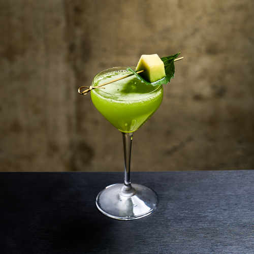 A bright green cocktail in a stemmed coupe glass garnished with a melon cube and a leaf on a bamboo pick against a textured brown wall.