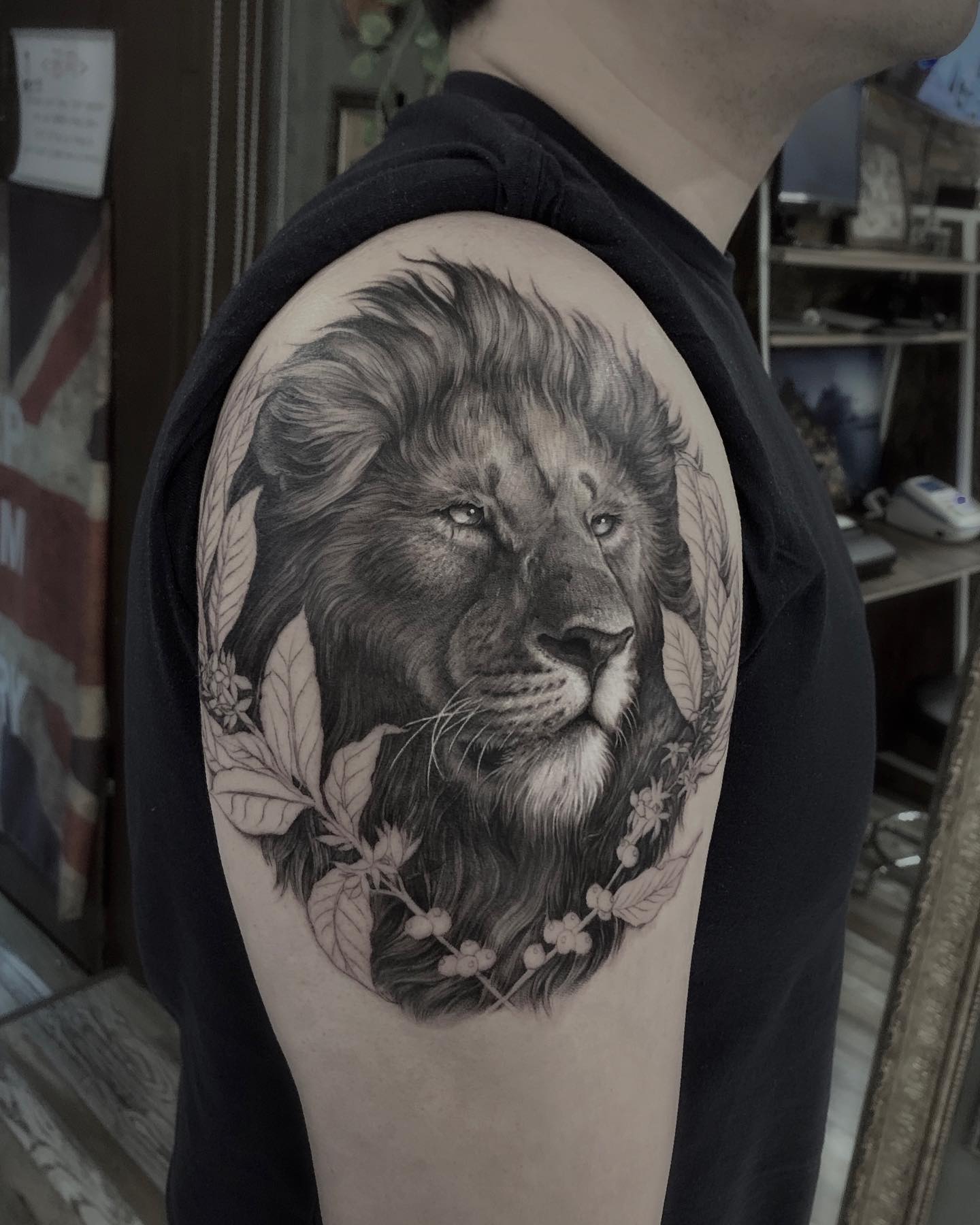A detailed black and grey realistic lion tattoo on a person's upper arm, surrounded by botanical leaf and berry motifs.