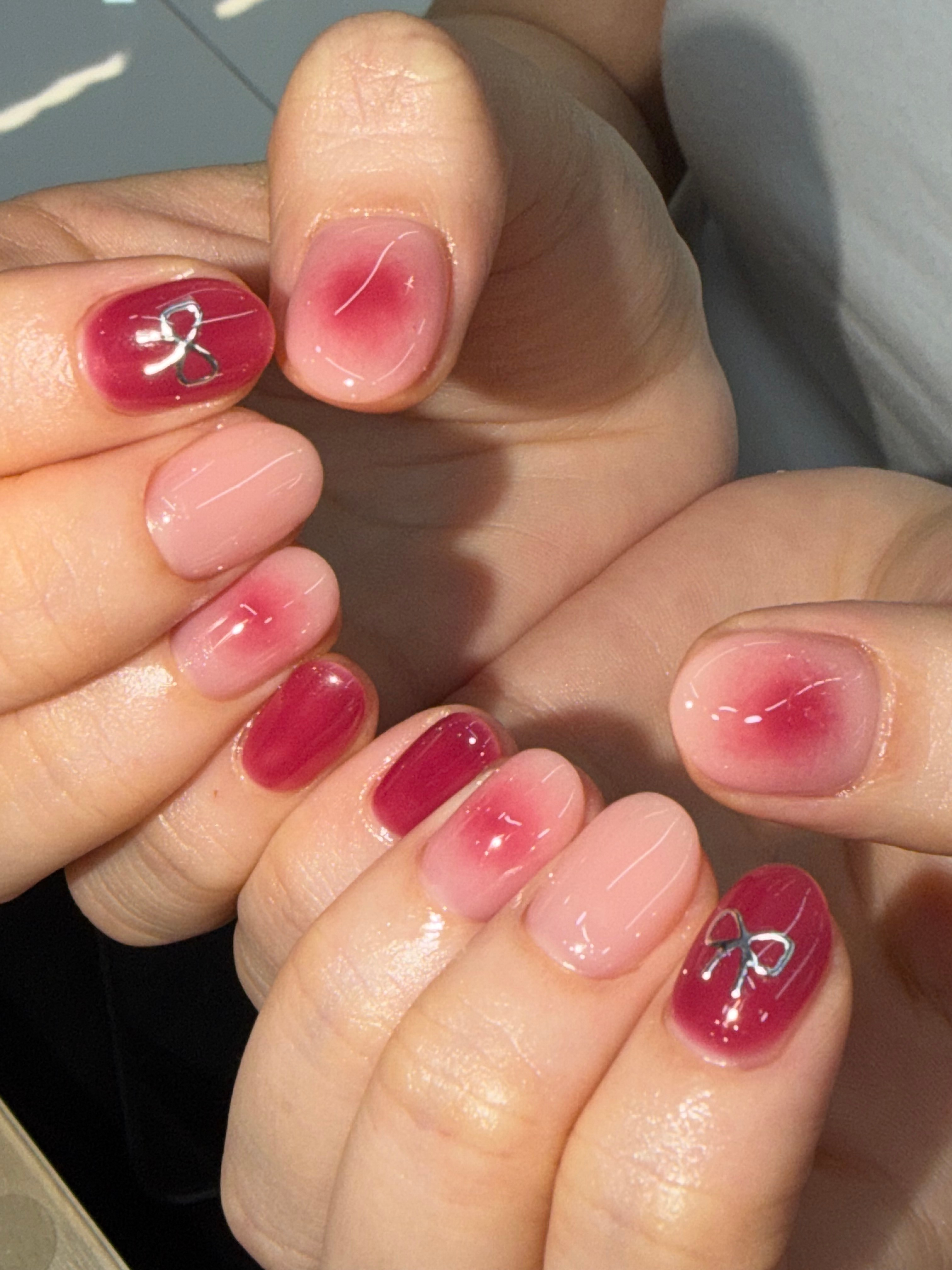 Close-up of hands with nail art featuring a mix of pink and red ombre polish and silver bow accents