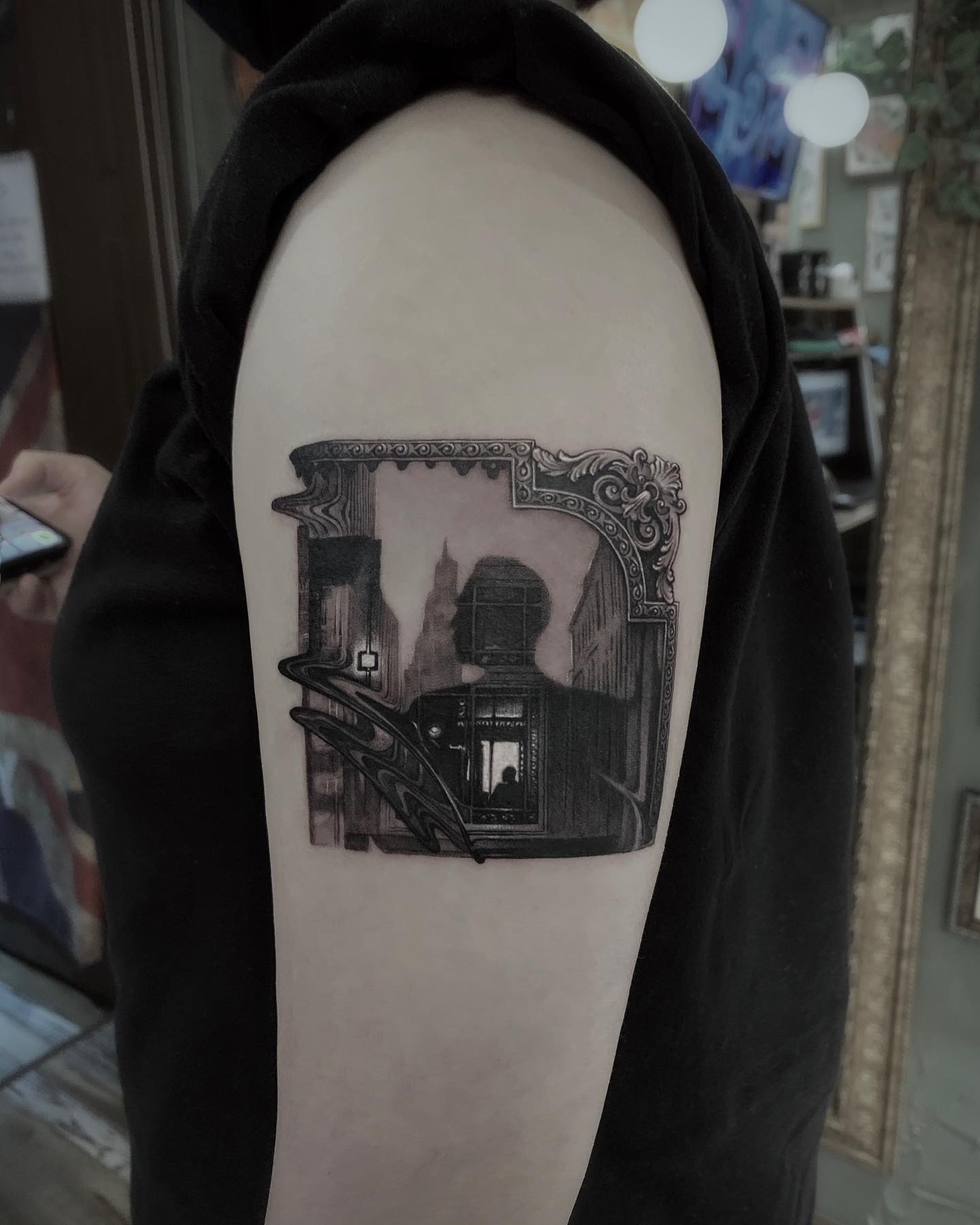 A detailed black and grey tattoo on an upper arm featuring a surreal scene framed by an ornate corner piece.