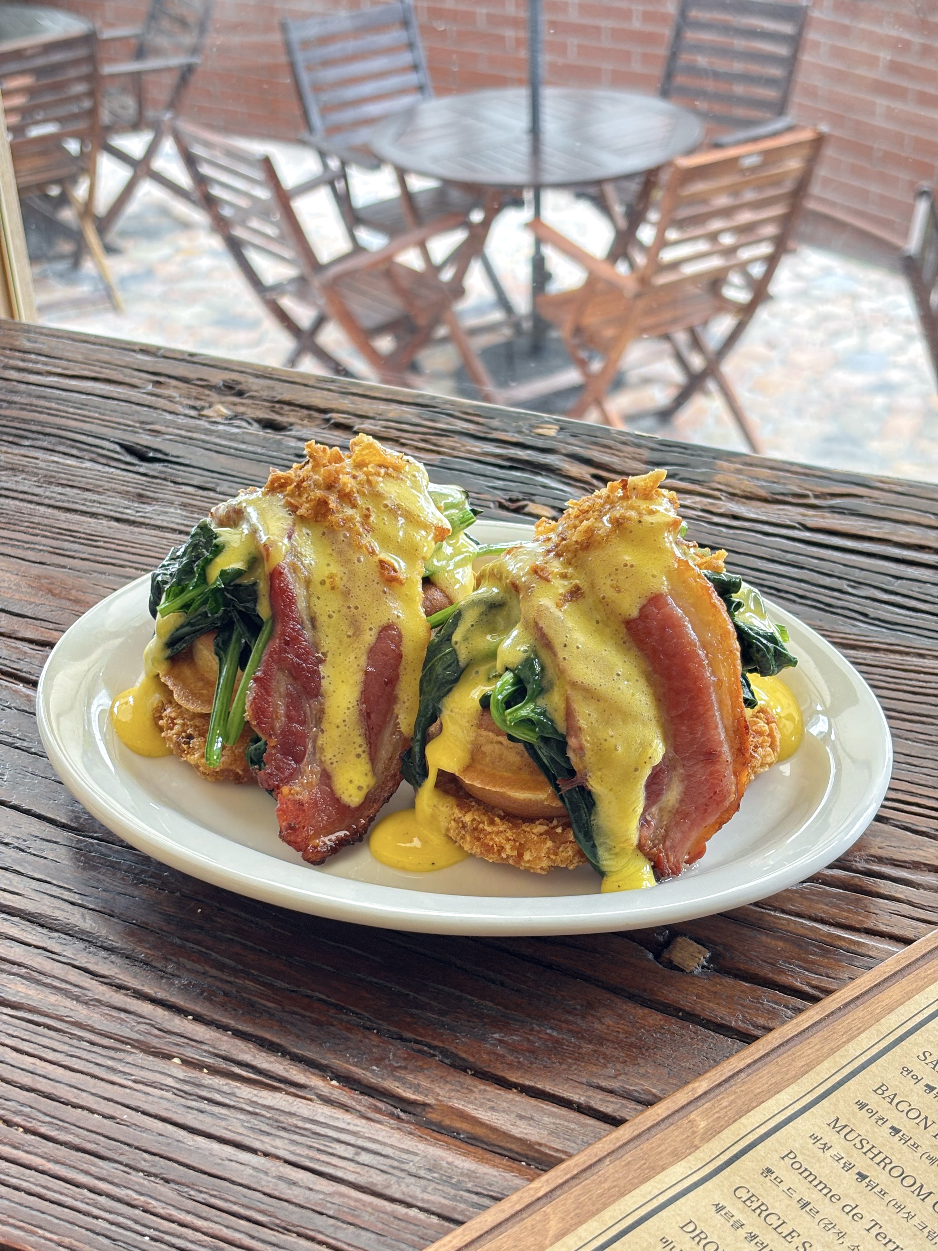 Two servings of brunch featuring waffles, bacon, greens, and yellow sauce on a white plate on a wooden table.