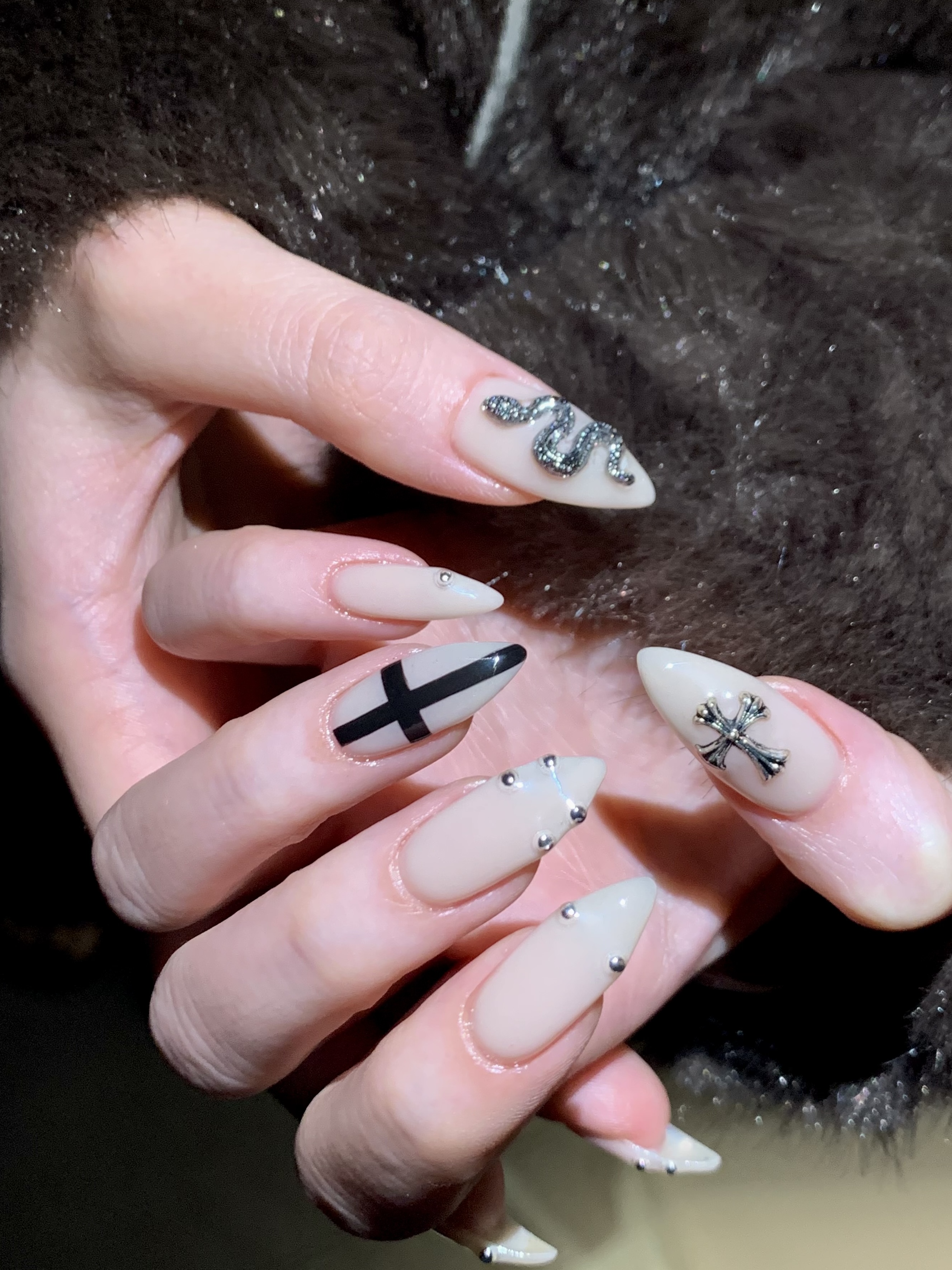 Close-up of almond-shaped nails with cream polish featuring snake charm, cross design, and small metallic studs