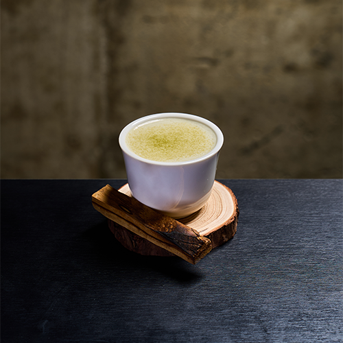 A white cup of light green liquid on a wooden coaster with a piece of charred wood, against a concrete-textured background.