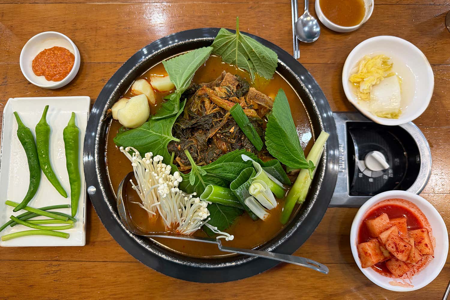 A flat-lay view of a traditional Gamjatang pork bone soup meal served in a black pot with side dishes on a wooden table.
