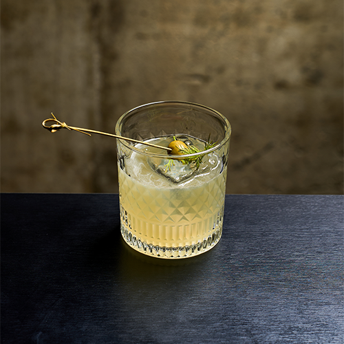 A crystal glass with a pale yellow cocktail garnished with an olive and a sprig of dill on a black table against a concrete wall.