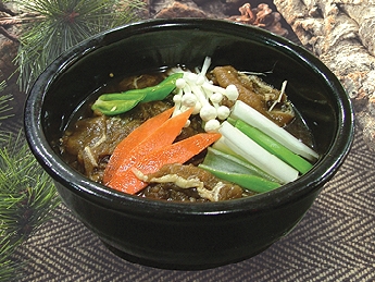 A close-up shot of a black bowl filled with Korean stew, garnished with enoki mushrooms, carrots, and green vegetables.