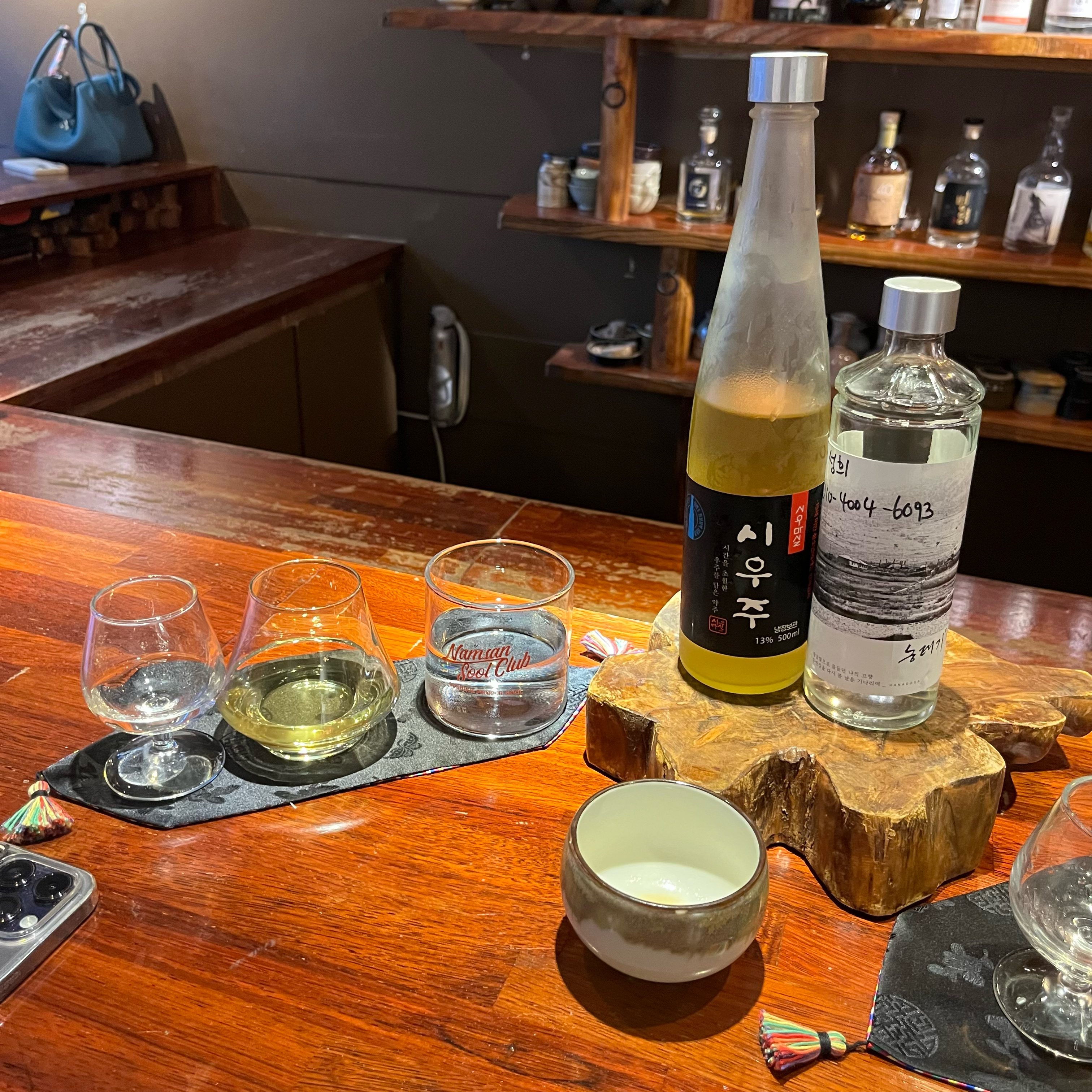 Two bottles of alcohol and assorted glassware on a wooden bar counter at Namsan Sool Club.