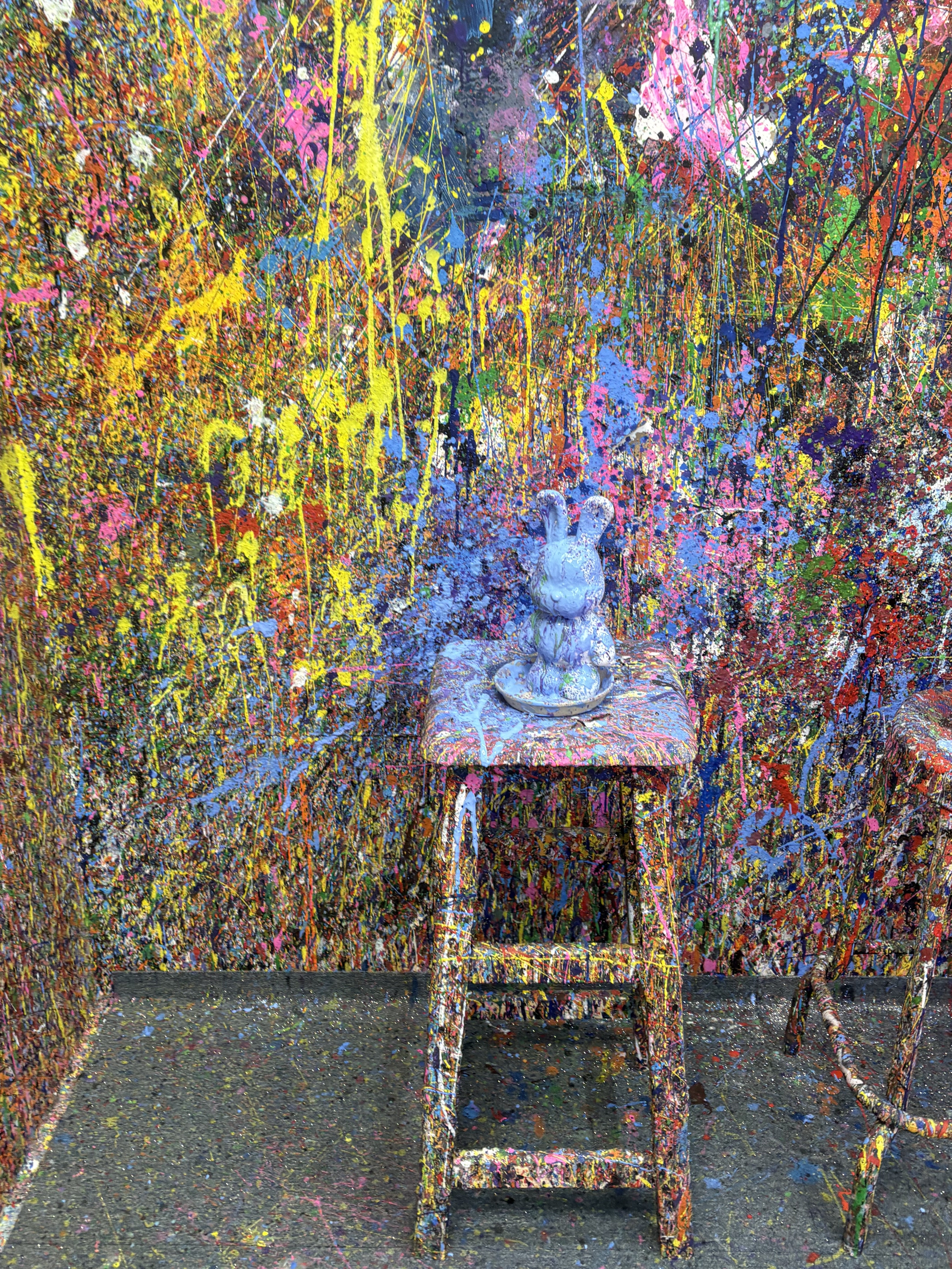A light blue rabbit figurine on a paint-splattered stool against a brightly colored, abstract paint-splattered wall
