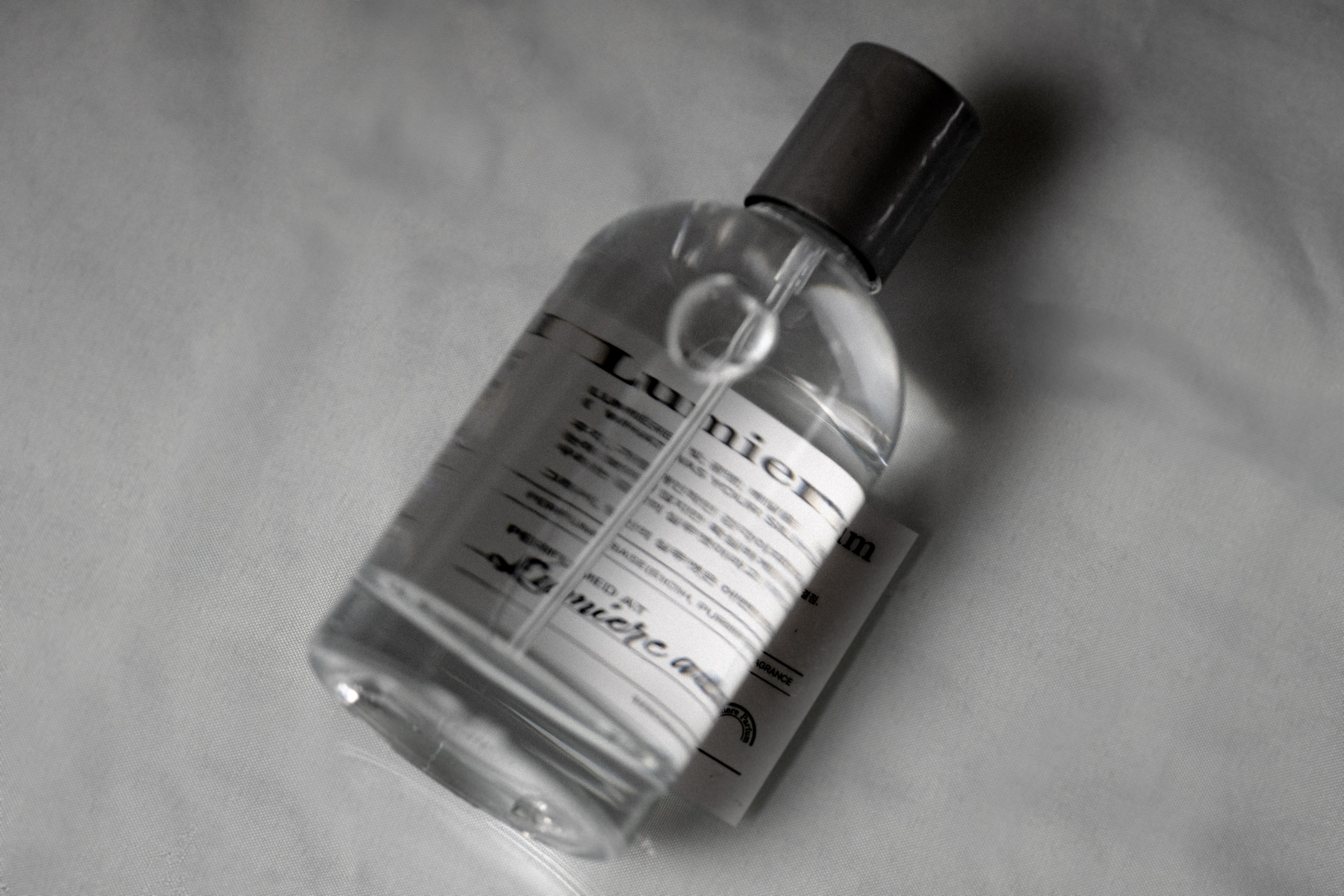 A clear glass perfume bottle with a dark cap sitting on a textured off-white fabric surface