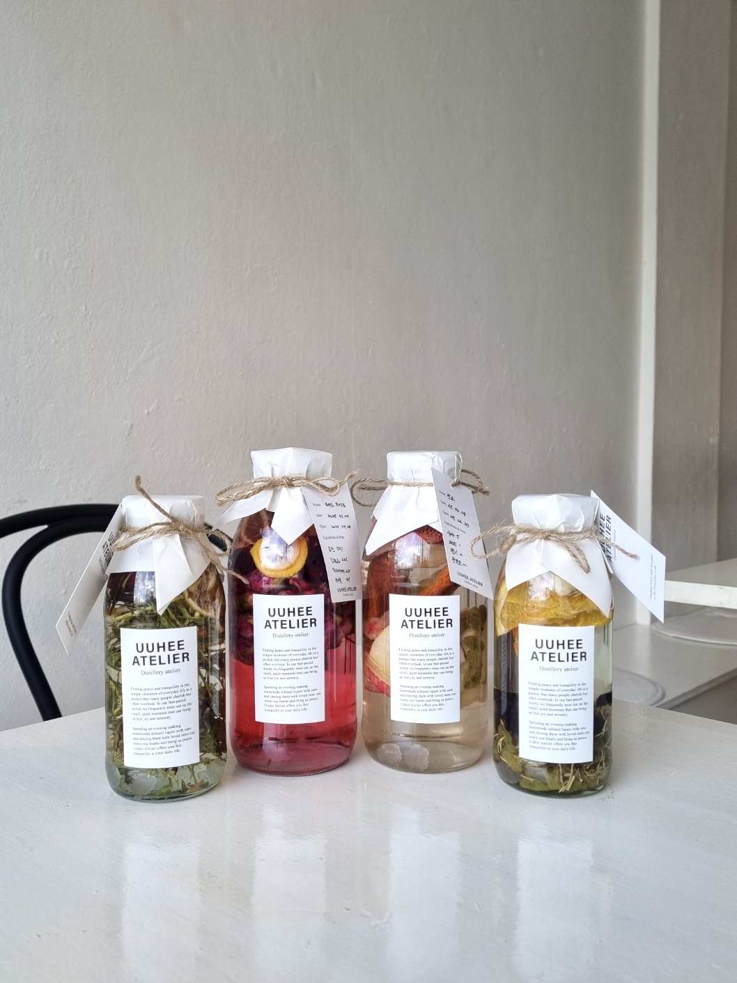 Four glass bottles filled with dried fruit, herbs, and liquid, labeled UUHEE ATELIER on a white table.
