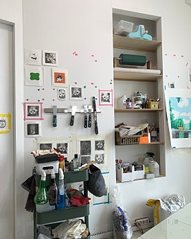 An art studio wall featuring prints, tools on a magnetic rack, and a shelving unit with supplies.