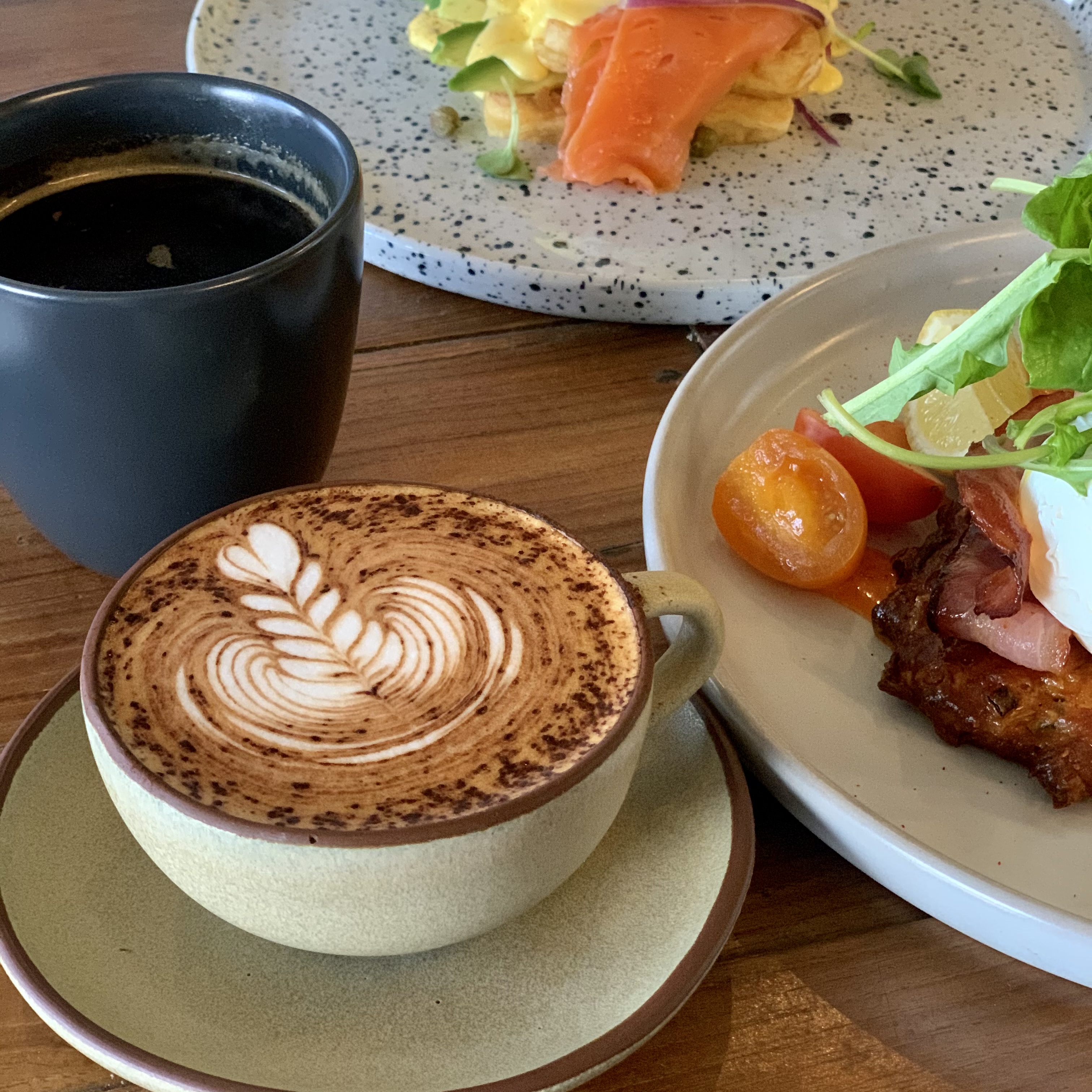 A flat lay of a latte with latte art, black coffee, and brunch plates on a wooden table.