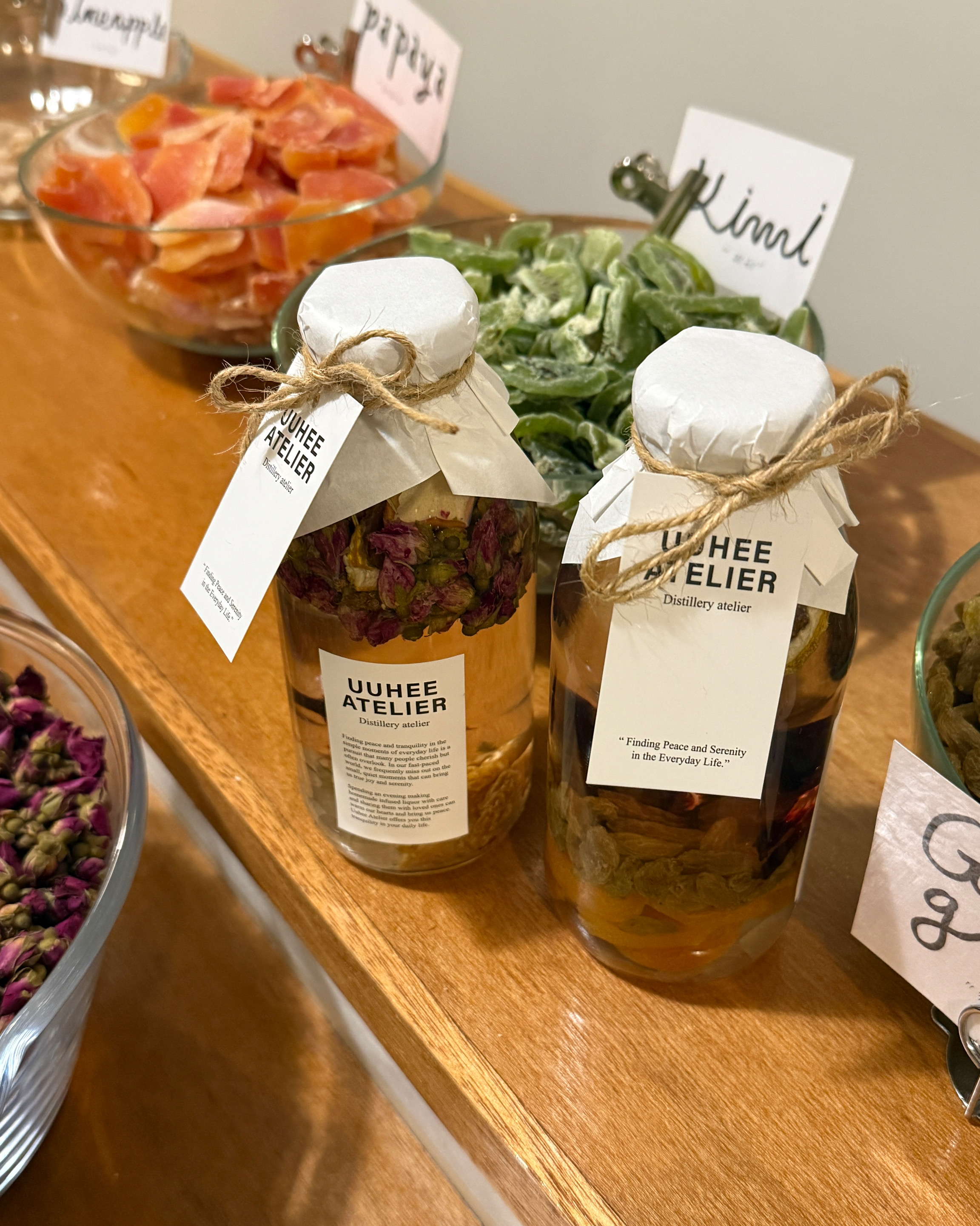 Two glass bottles of infused liquor with tags on a wooden shelf in a craft atelier.