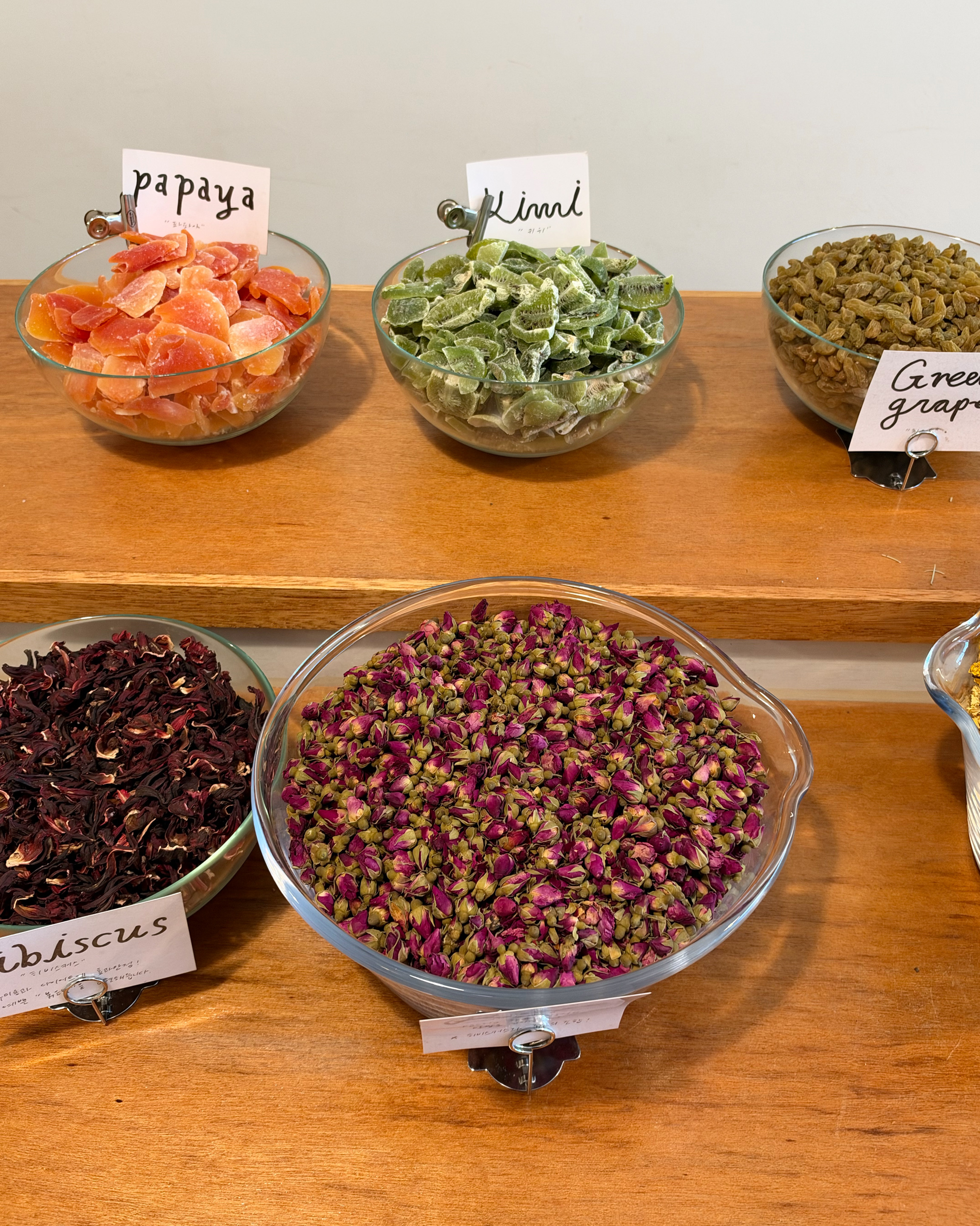 Five glass bowls filled with dried fruits and flowers displayed on wooden shelves at a workshop.