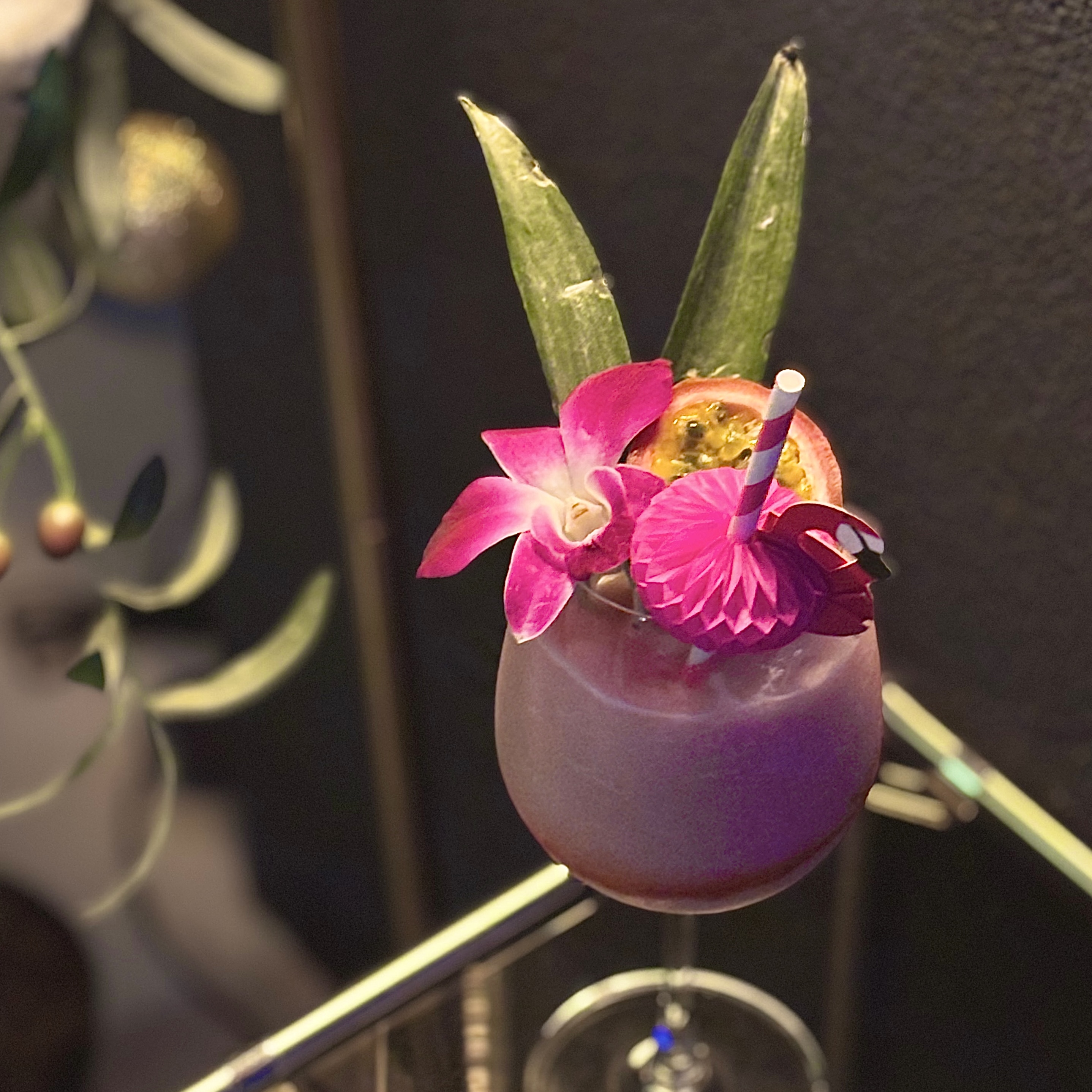 Pink cocktail garnished with purple orchid, pineapple leaves, and passionfruit