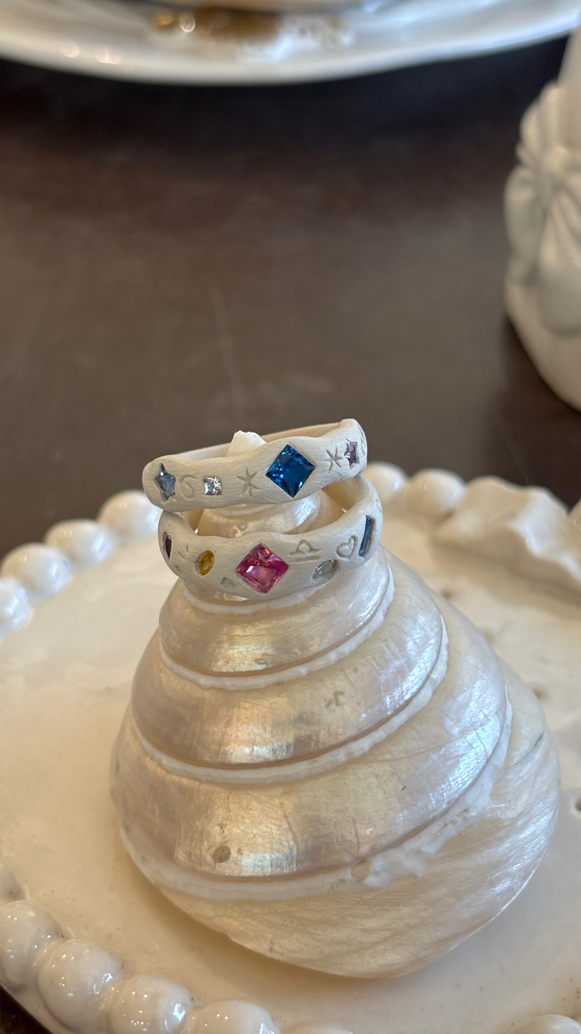 Two handmade textured rings with colorful gemstones stacked on a spiral seashell display.