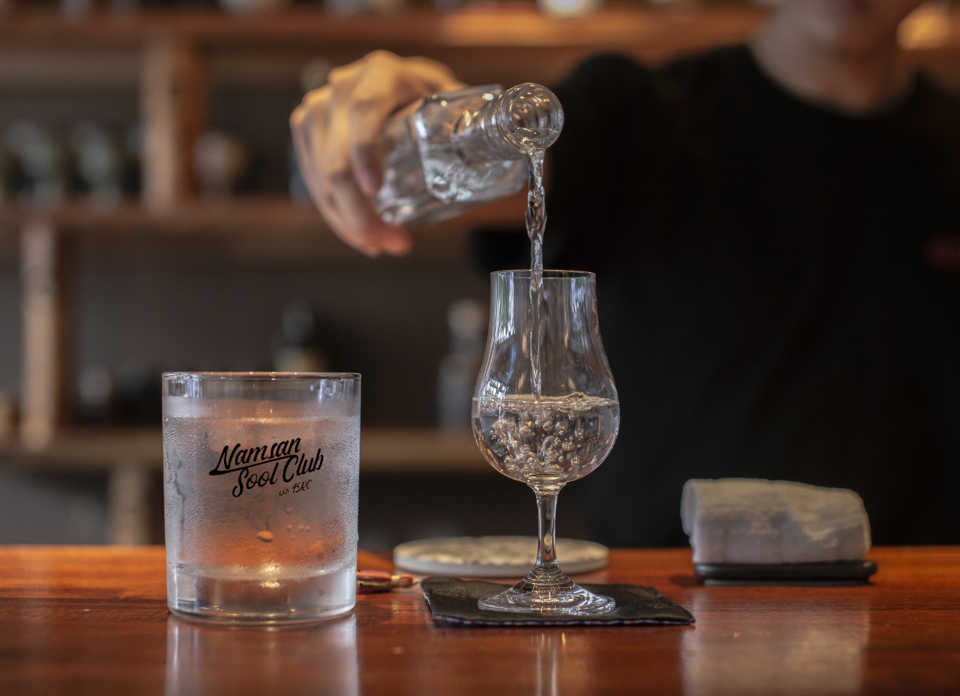 A bartender pours a clear spirit from a bottle into a tulip-shaped glass on a wooden bar counter.