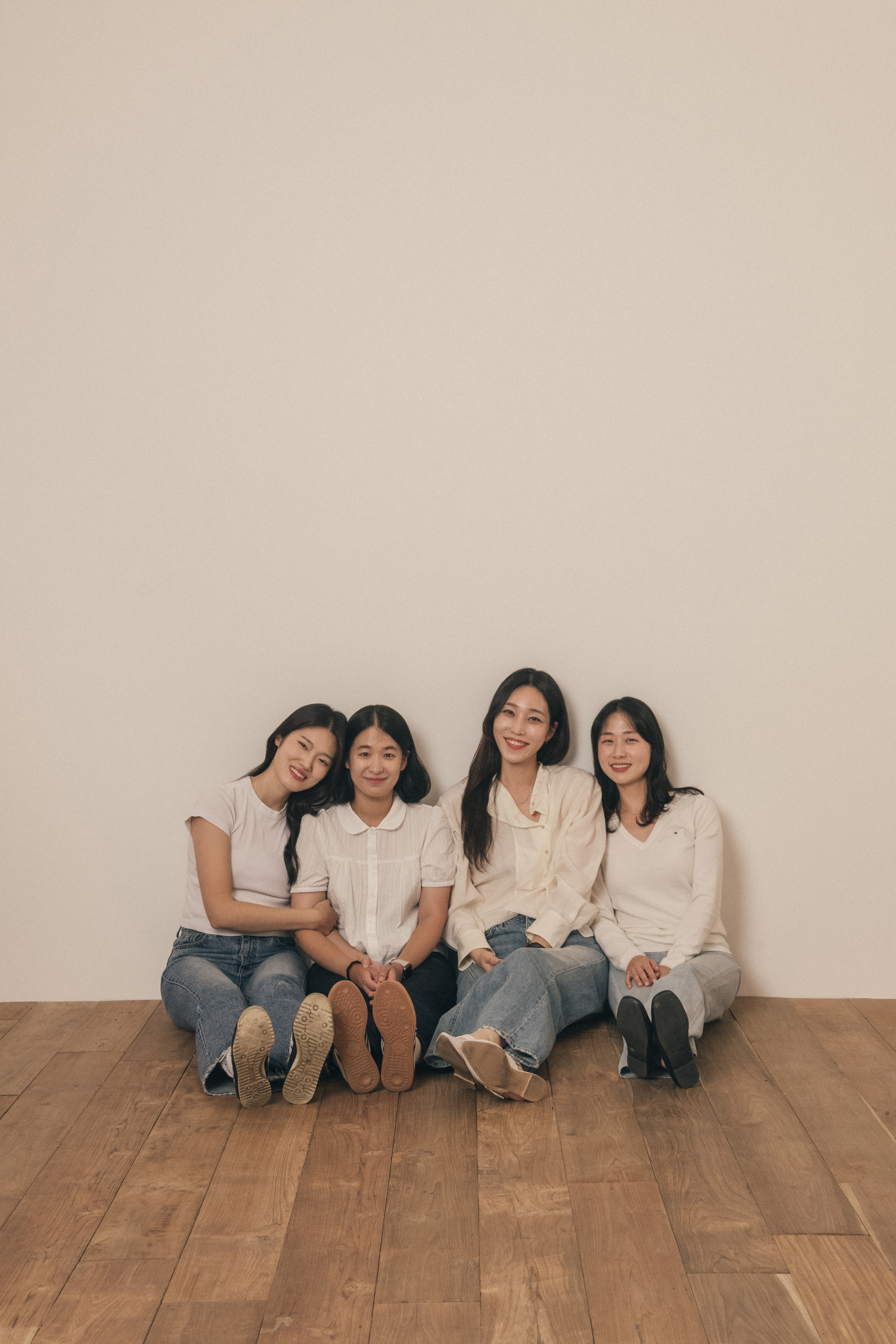 Four women sitting in a row against a plain beige studio wall on a wooden floor