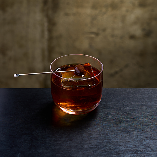 An amber cocktail in a lowball glass with a large clear ice cube and a cocktail pick on a dark wooden table.