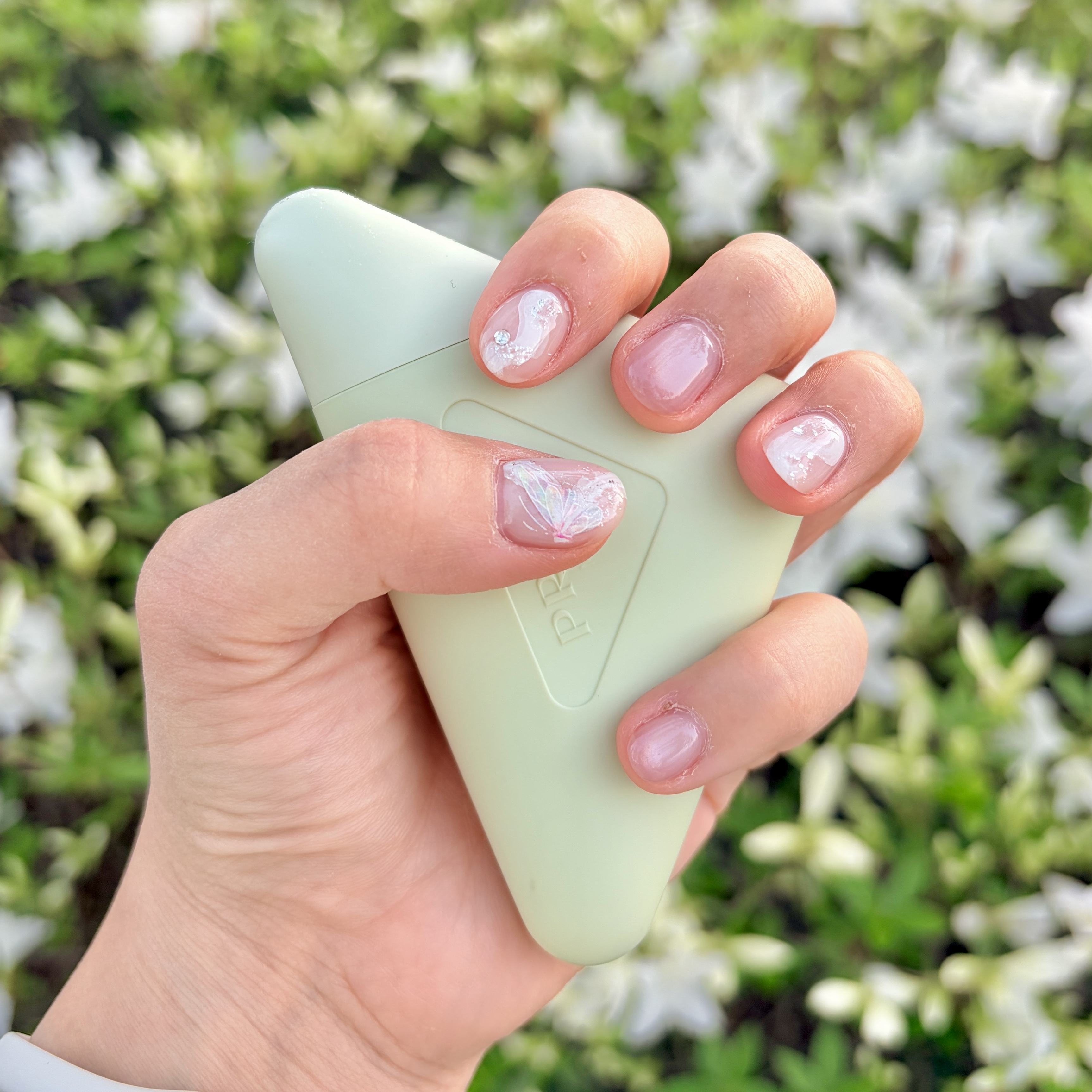 Hand with decorated nails holding pale green object featuring iridescent butterfly wings and rhinestones