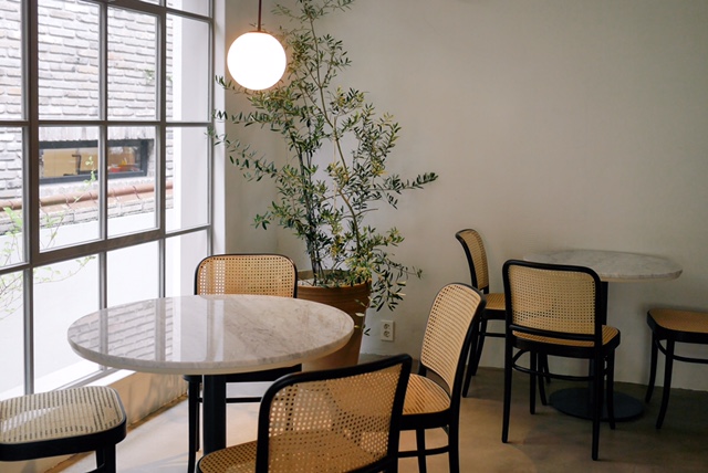 Interior of Oasis Hannam cafe with marble bistro tables, cane chairs, and a large potted tree near a window