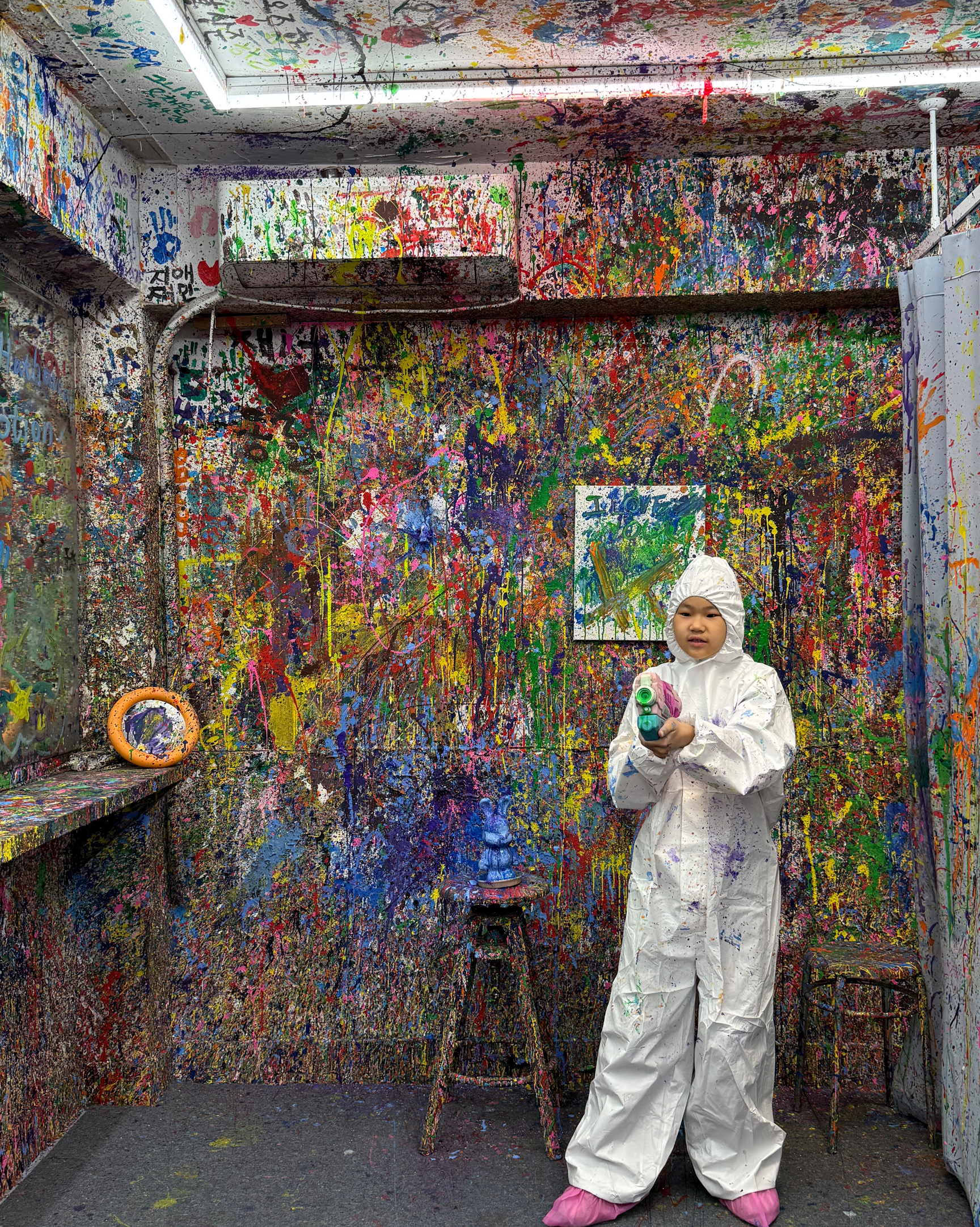 A person in a white protective suit holding a toy paint gun in a studio with walls covered in colorful paint splashes