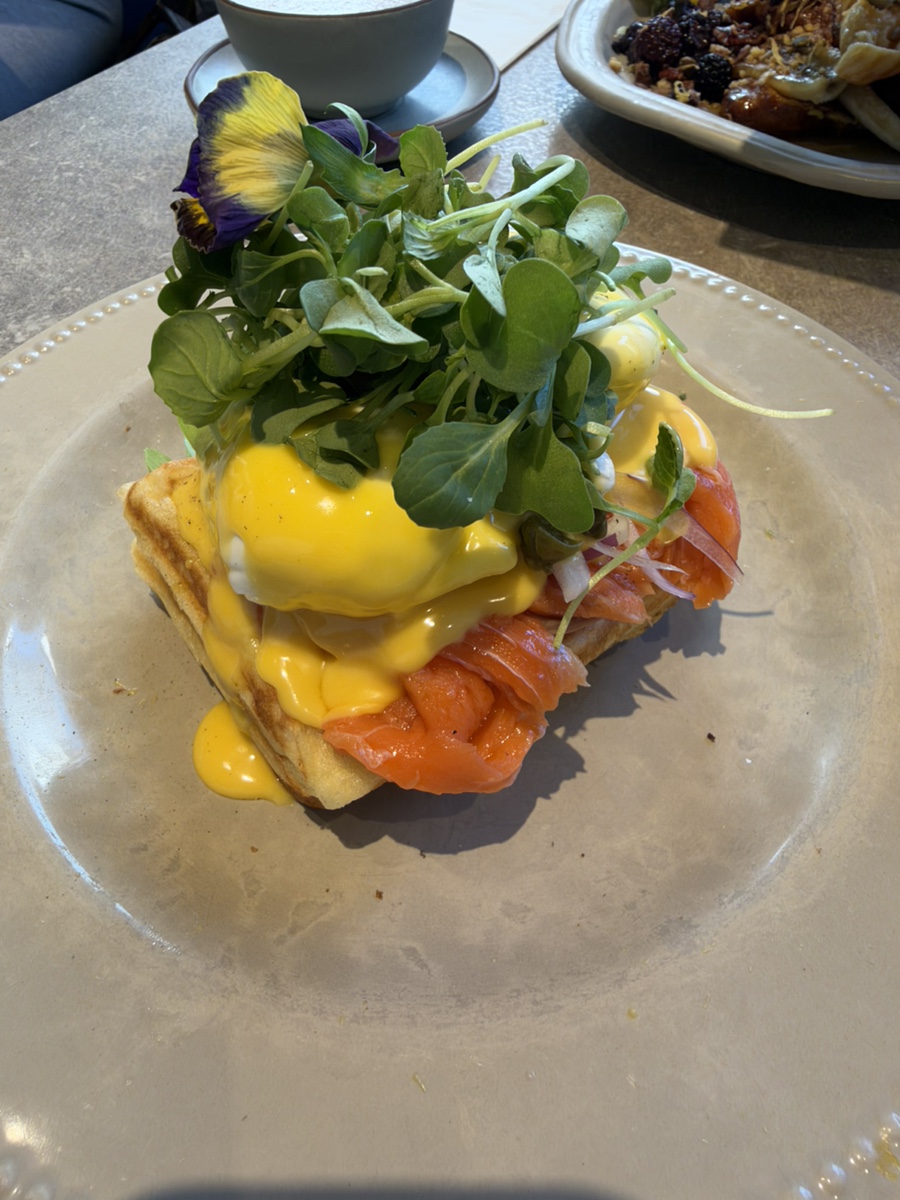 A waffle topped with smoked salmon, poached eggs, hollandaise sauce, and fresh greens on a plate.