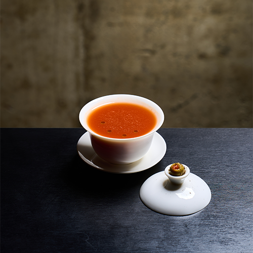 An orange cocktail served in a white tea cup with its lid placed nearby on a dark surface against a textured concrete wall.