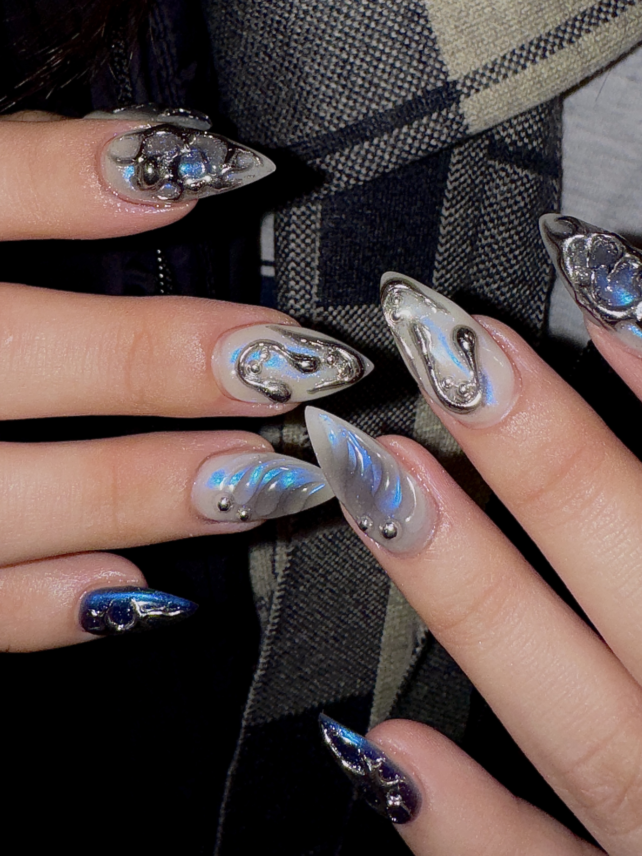 Close-up of stiletto-shaped nails with chrome 3D art, iridescent blue accents, and metallic spheres against plaid fabric