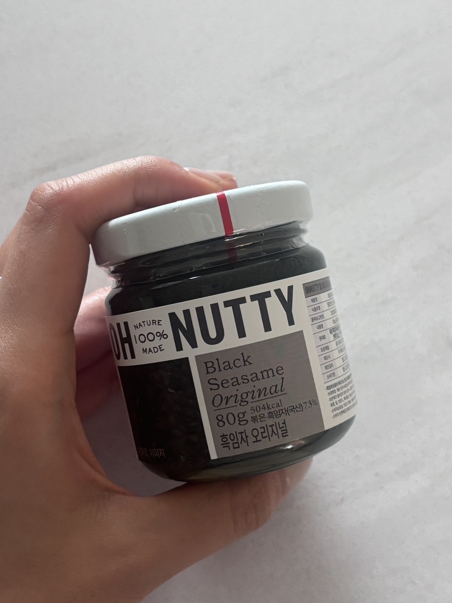 A hand holding a small glass jar of Oh Nutty Black Sesame spread against a light gray background.