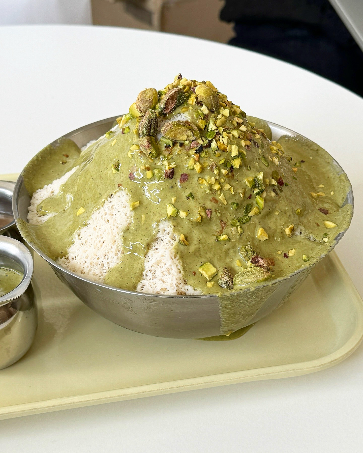 A bowl of shaved ice dessert topped with green pistachio sauce and chopped pistachios on a tray in a cafe.