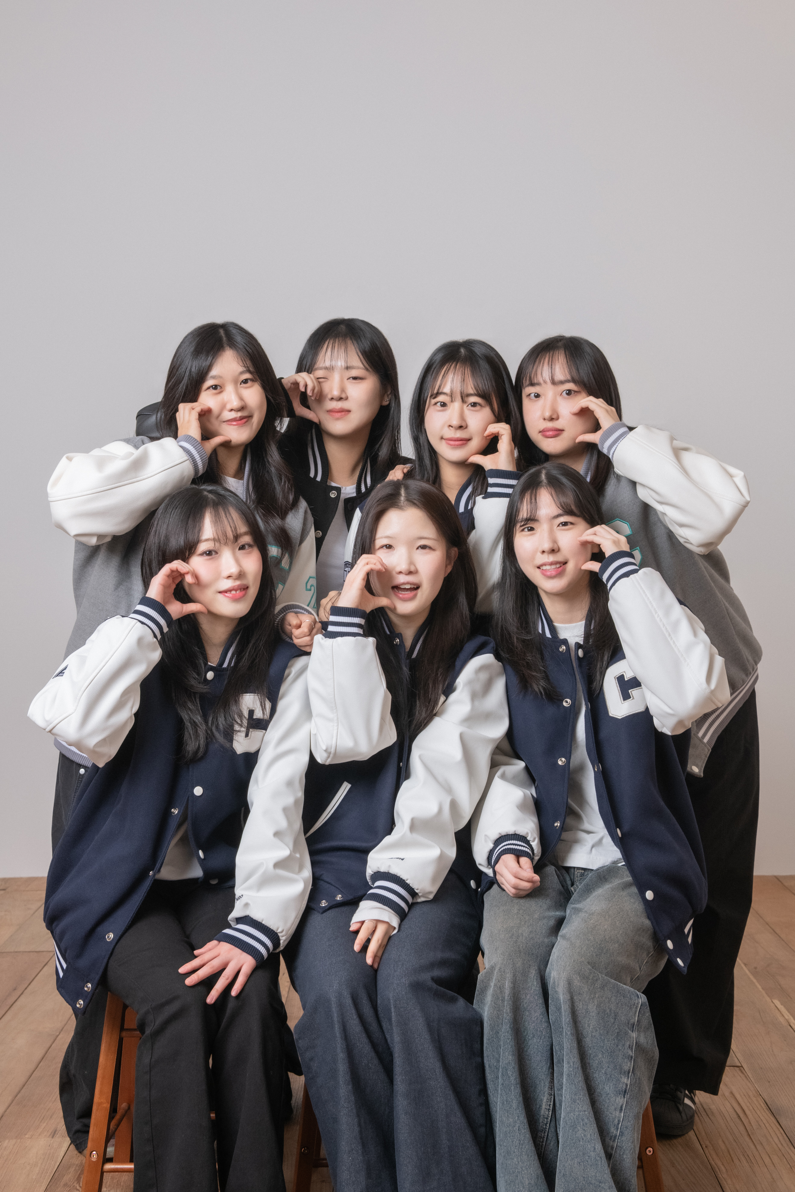Seven young women posing together wearing matching varsity jackets against a plain grey backdrop in a studio