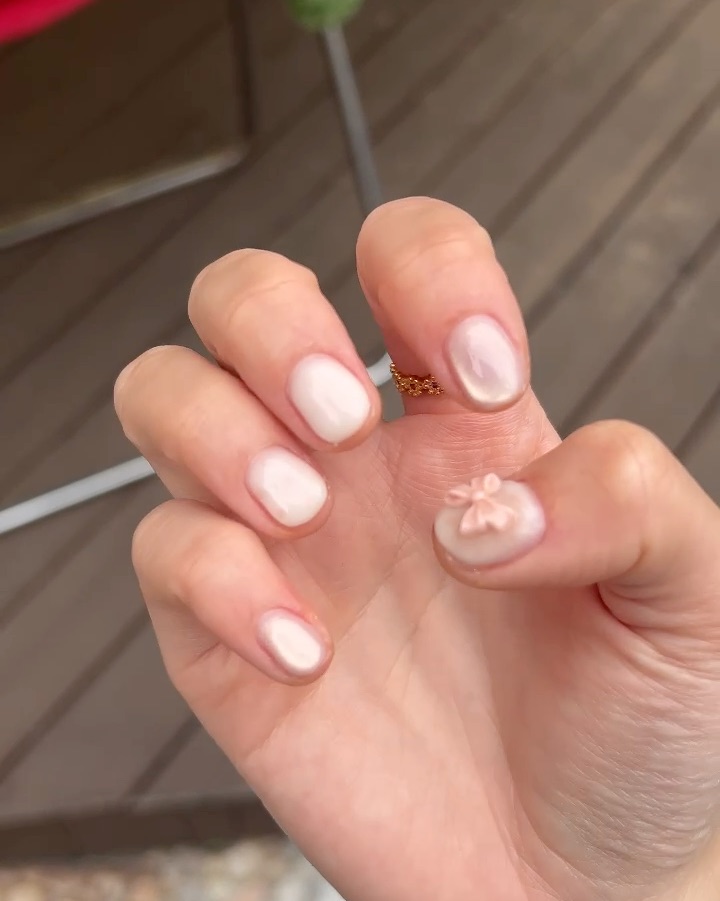 Close-up of a hand with short, glossy pearl-toned manicured nails and a small bow charm on the thumbnail