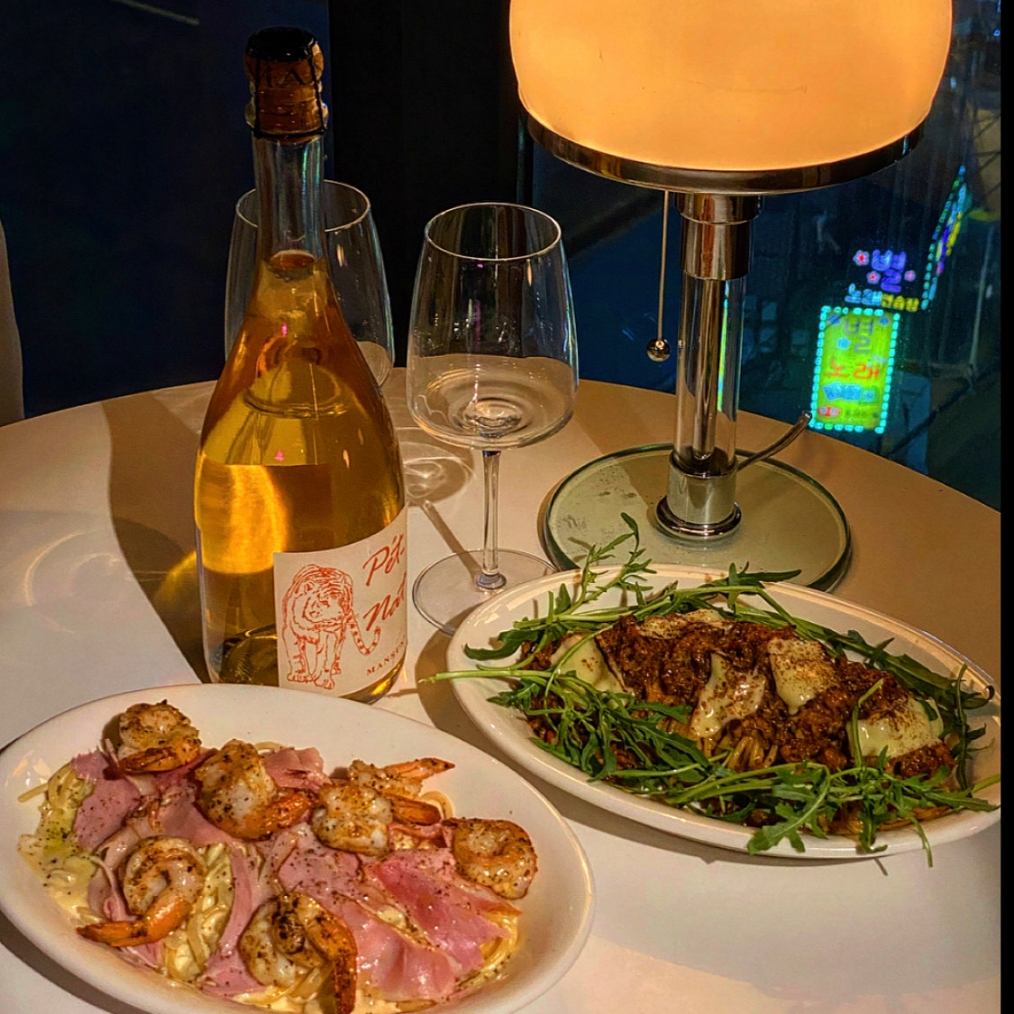 Pet-Nat wine bottle, pasta dishes, and glowing modern lamp on a white table