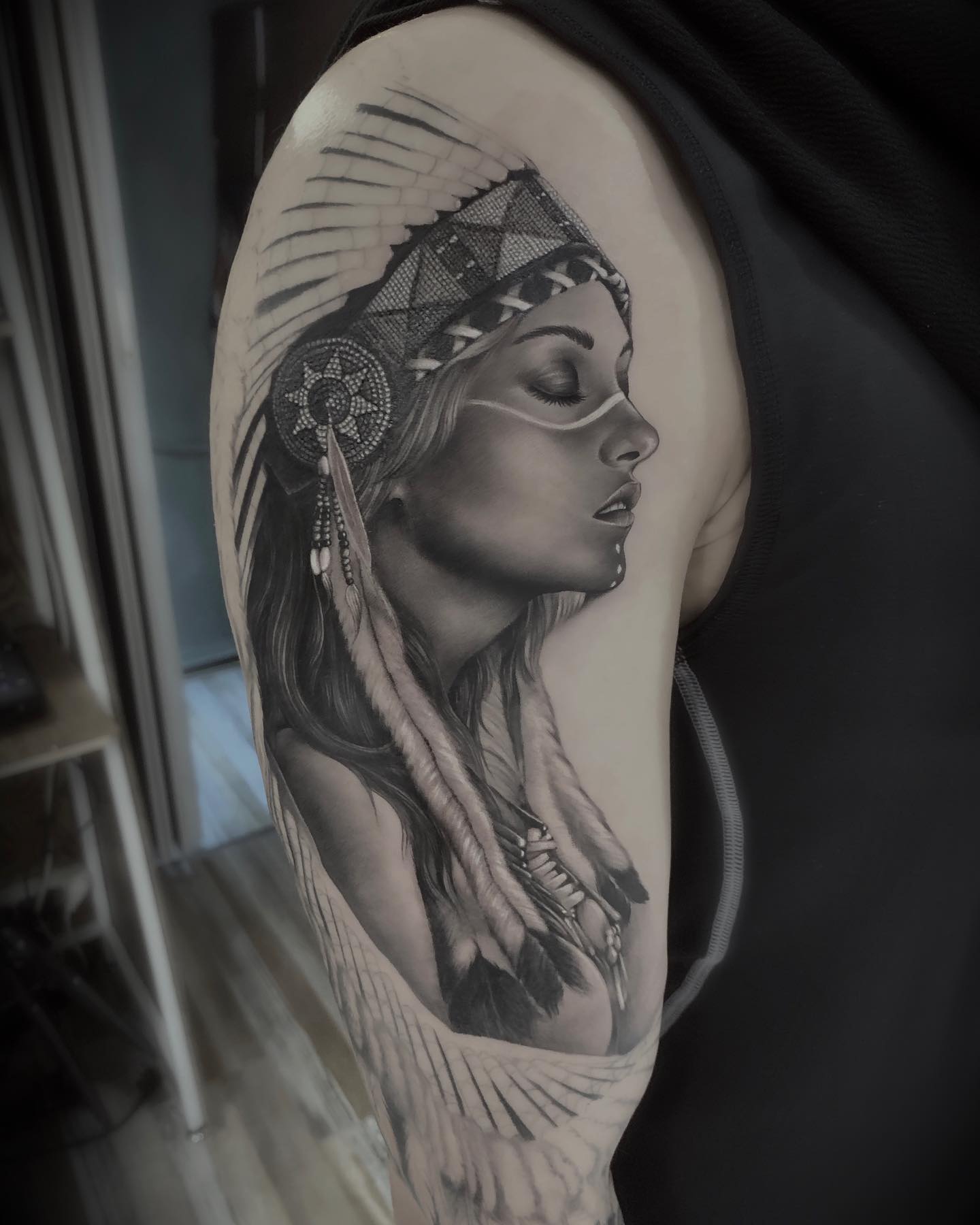 A detailed black and grey realistic tattoo of a woman wearing a Native American-style headdress on a person's upper arm.