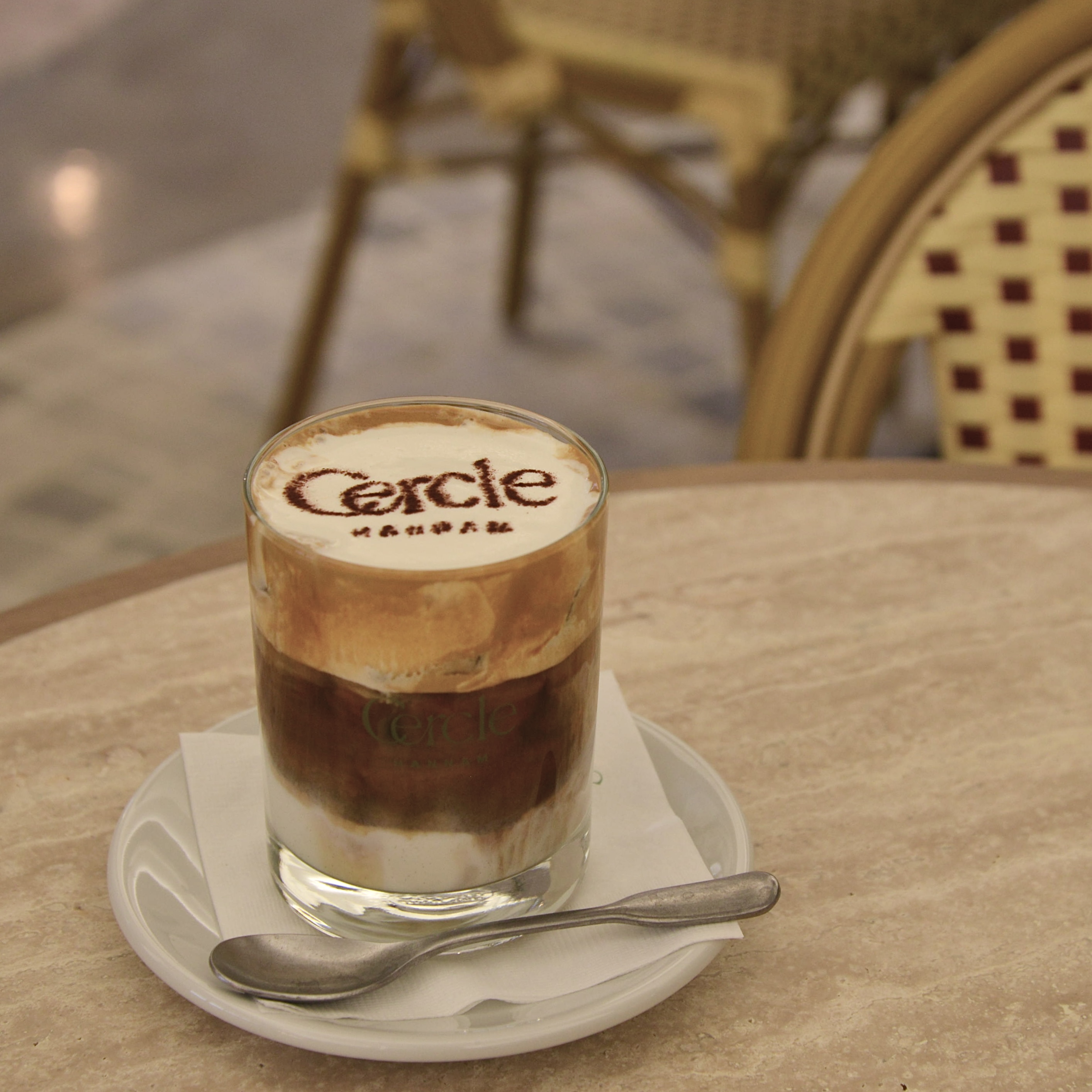 A layered coffee drink in a glass with cocoa powder branding, served on a marble table.
