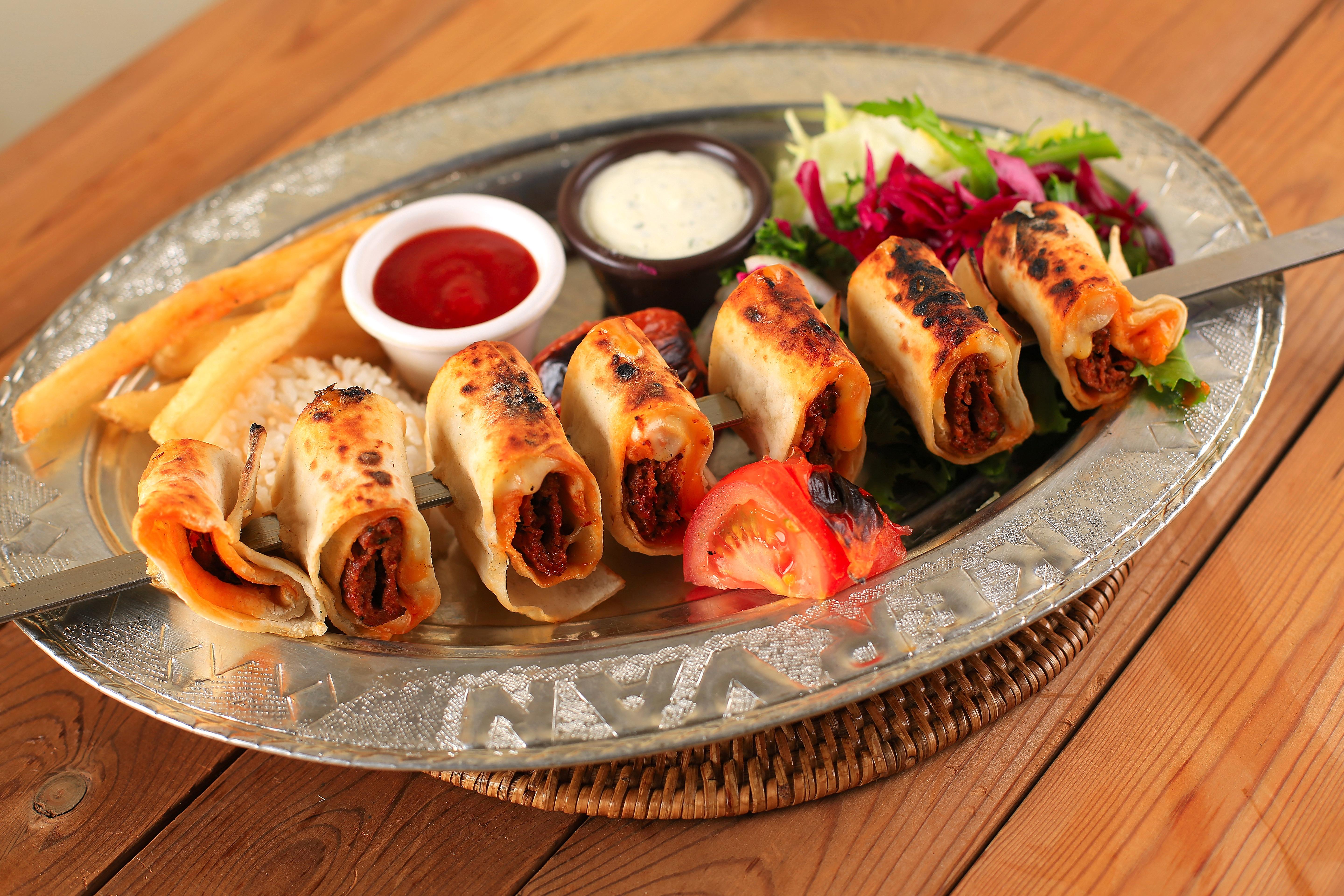 A metal platter featuring skewered meat wrapped in flatbread, served with fries, rice, salad, and dipping sauces.