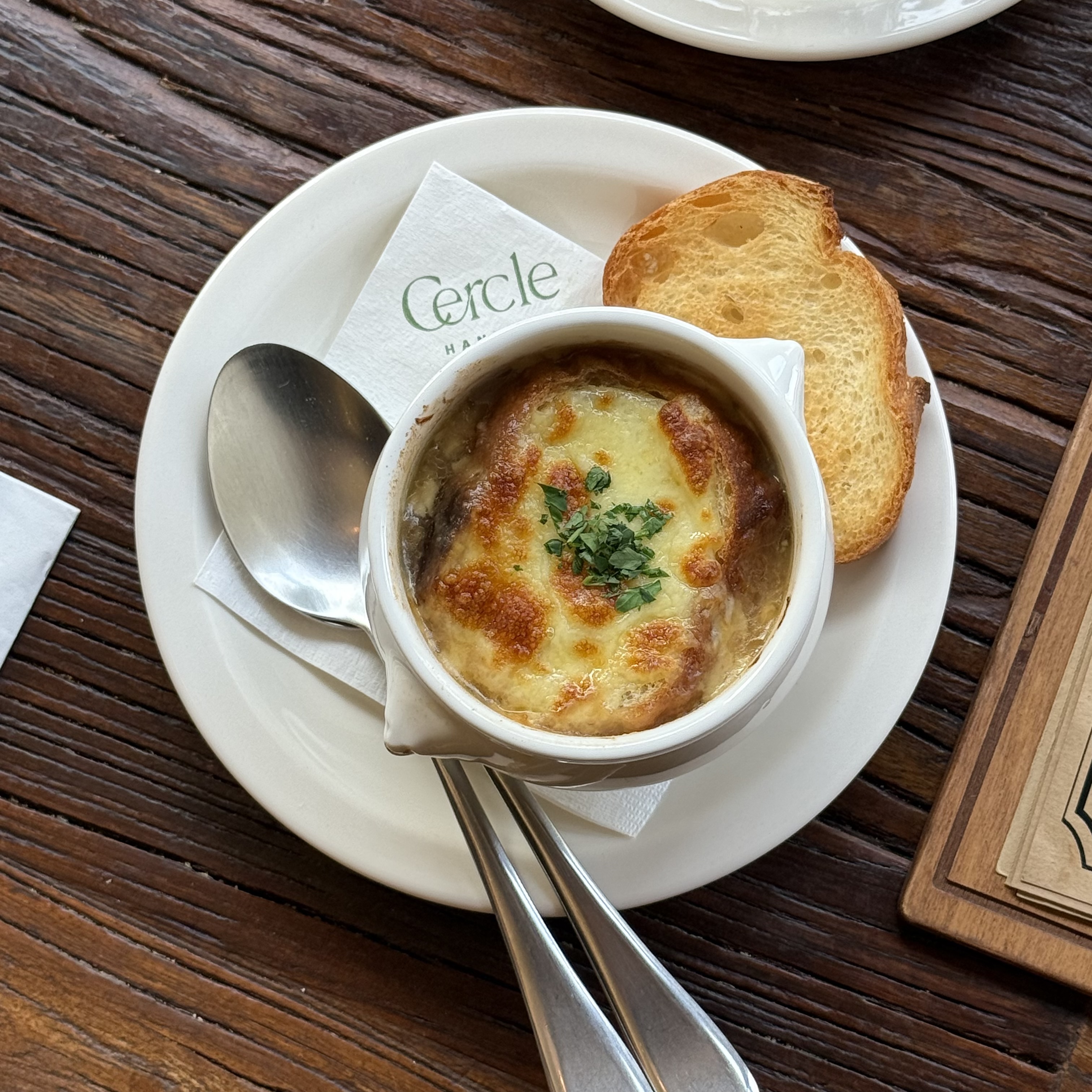 A bowl of onion soup topped with melted cheese and herbs, served with toasted bread on a branded napkin.
