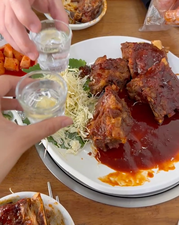 Spicy charcoal grilled pork spine dish served with shredded cabbage and soju glasses held in a toast.