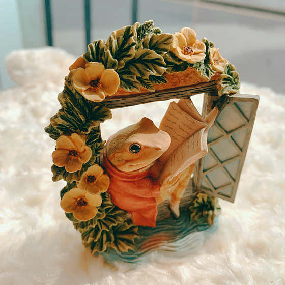 Resin frog figurine wearing a red vest and reading inside a floral window frame