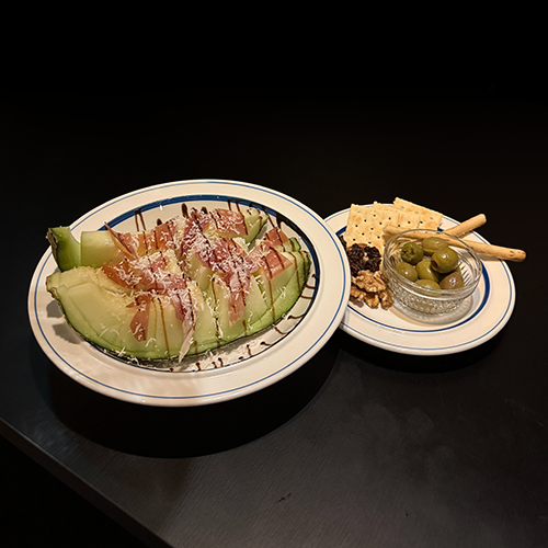 A plate of melon with prosciutto and a side of olives, crackers, and nuts against a dark background.