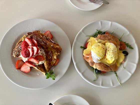 Two plates of brunch, one with strawberry French toast and one with eggs benedict, on a white table