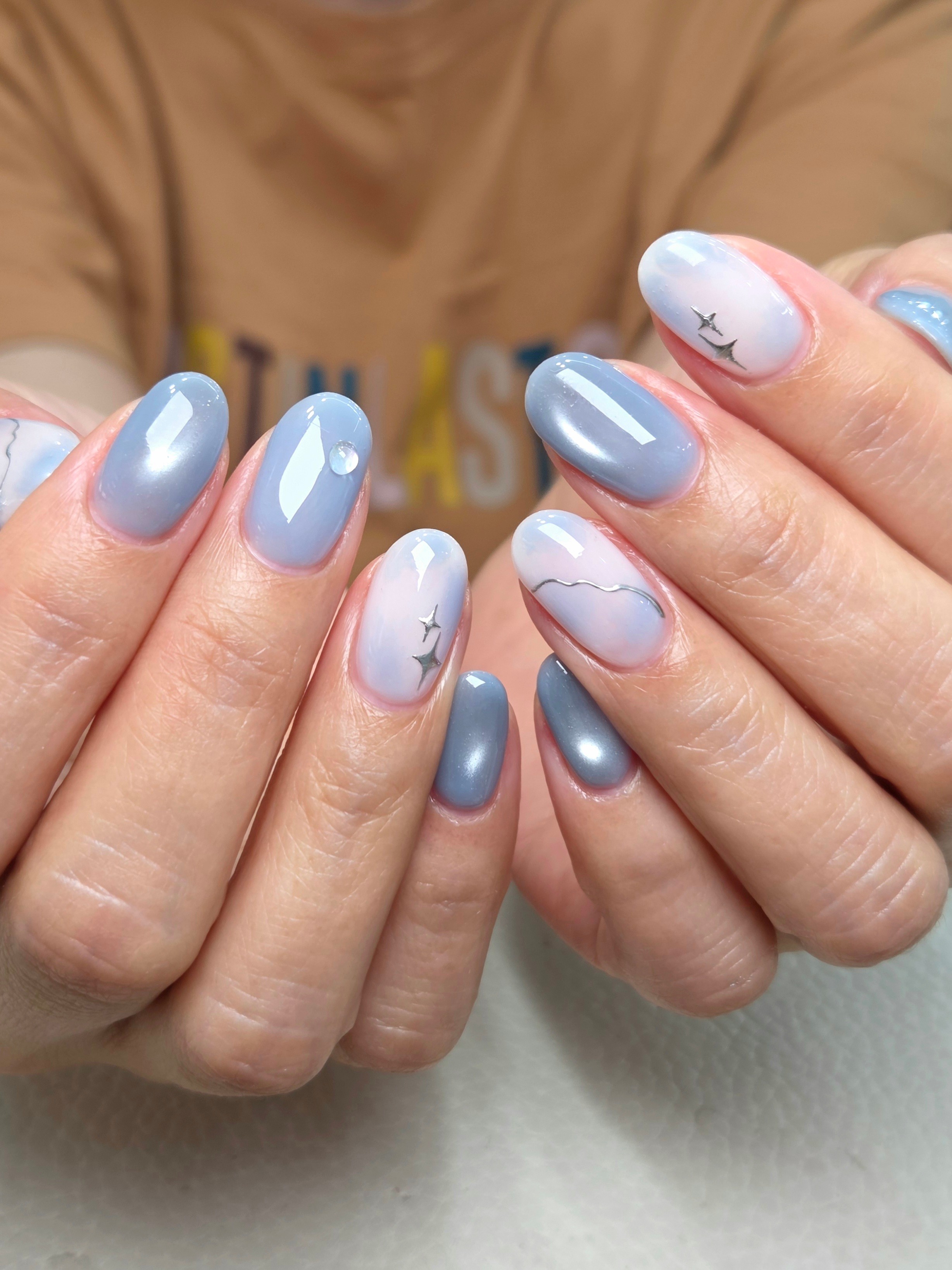 Close-up of manicured oval nails featuring blue and white gradient polish, star decals, and metallic accents
