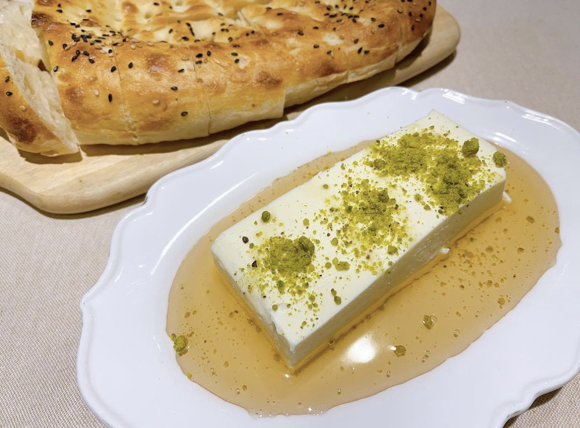 A rectangular block of white kaymak cream topped with green pistachio crumbles, served with honey and flatbread.