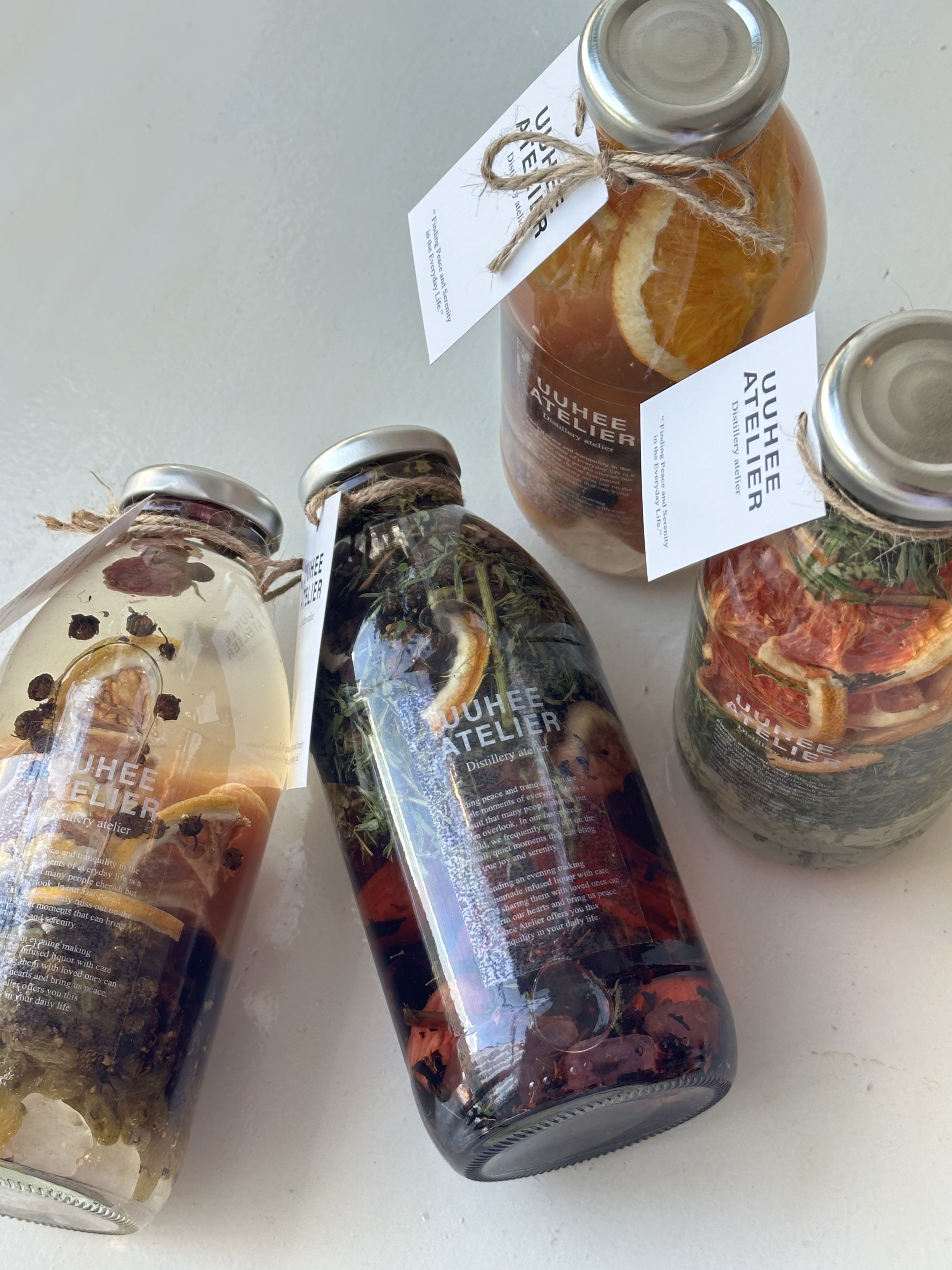Three glass bottles filled with fruit-infused liquid and herbs, each with a tag reading UUHEE ATELIER.