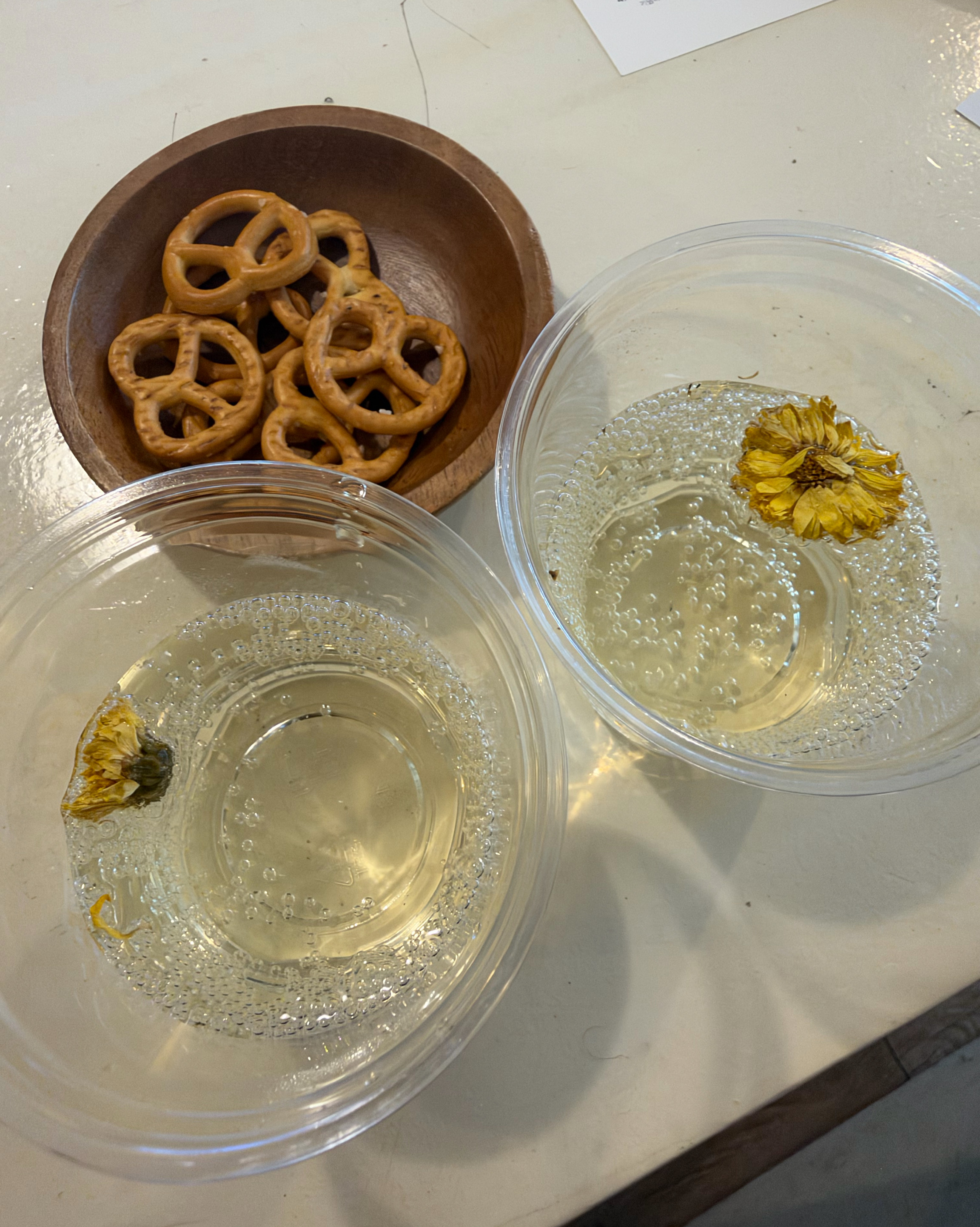 Two clear bowls of carbonated liquid with dried flowers and a wooden bowl of pretzels on a white table.
