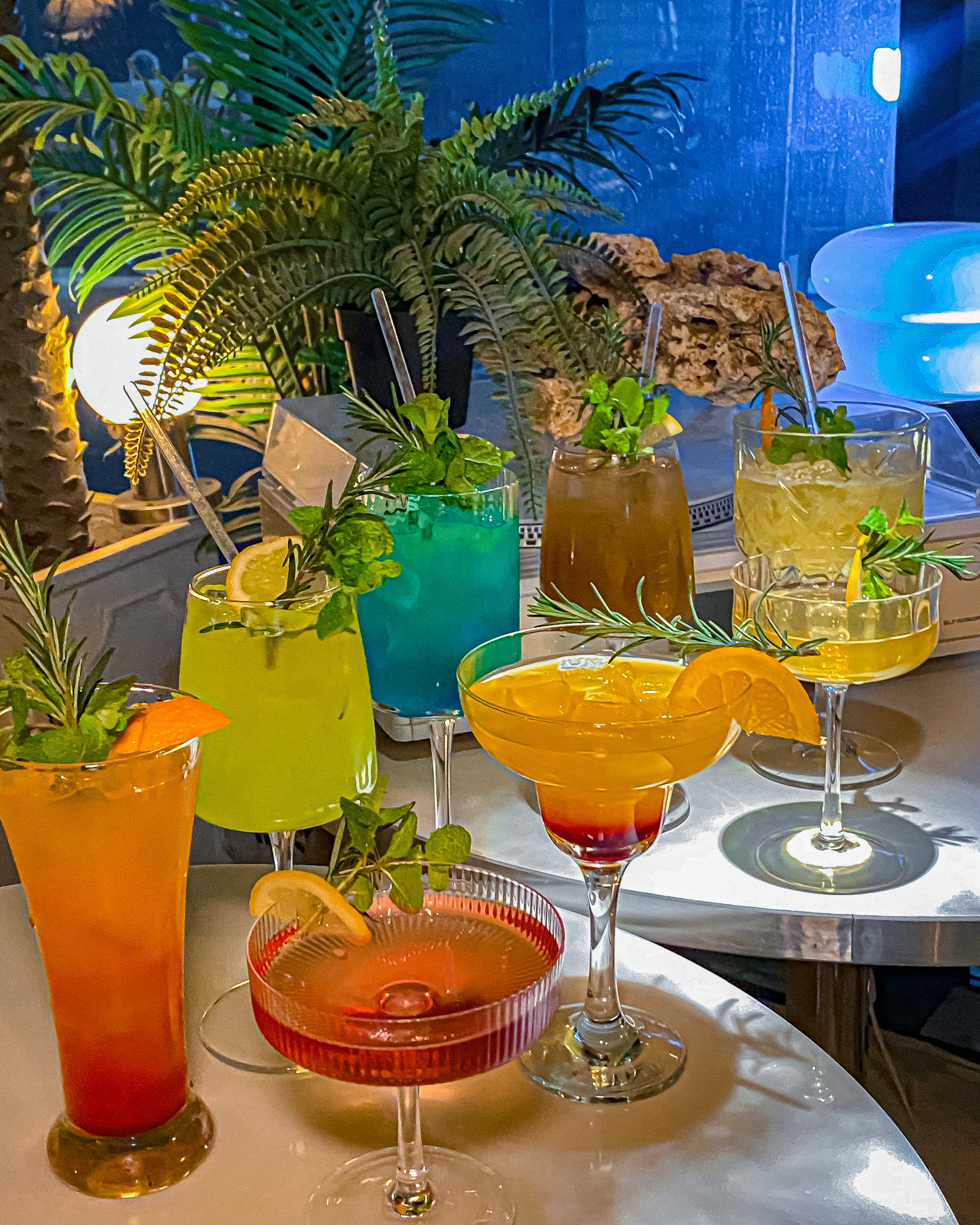 Cocktails in diverse glasses garnished with herbs and citrus on a metallic table