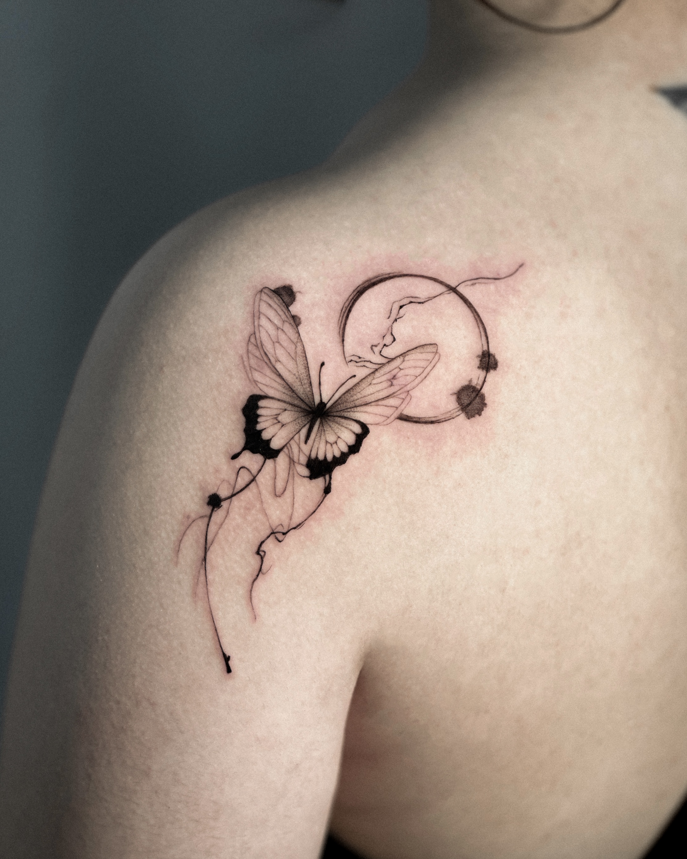 Delicate fine-line butterfly tattoo with abstract circular and ink-wash elements on shoulder