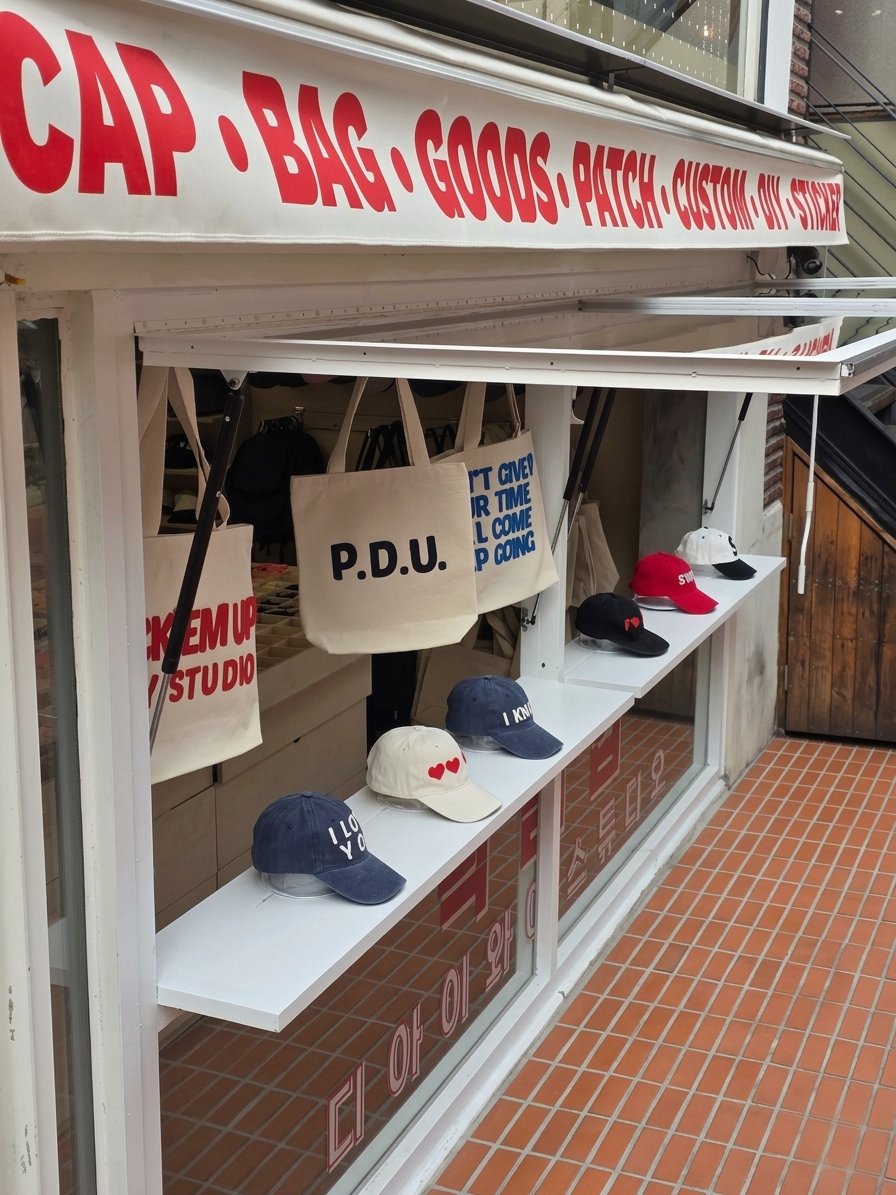 Storefront awning and window display of Pick'em Up studio with hanging tote bags and baseball caps