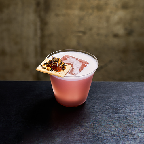A pink cocktail with a large ice cube, garnished with a small cracker topped with jam, set against a dark textured wall.